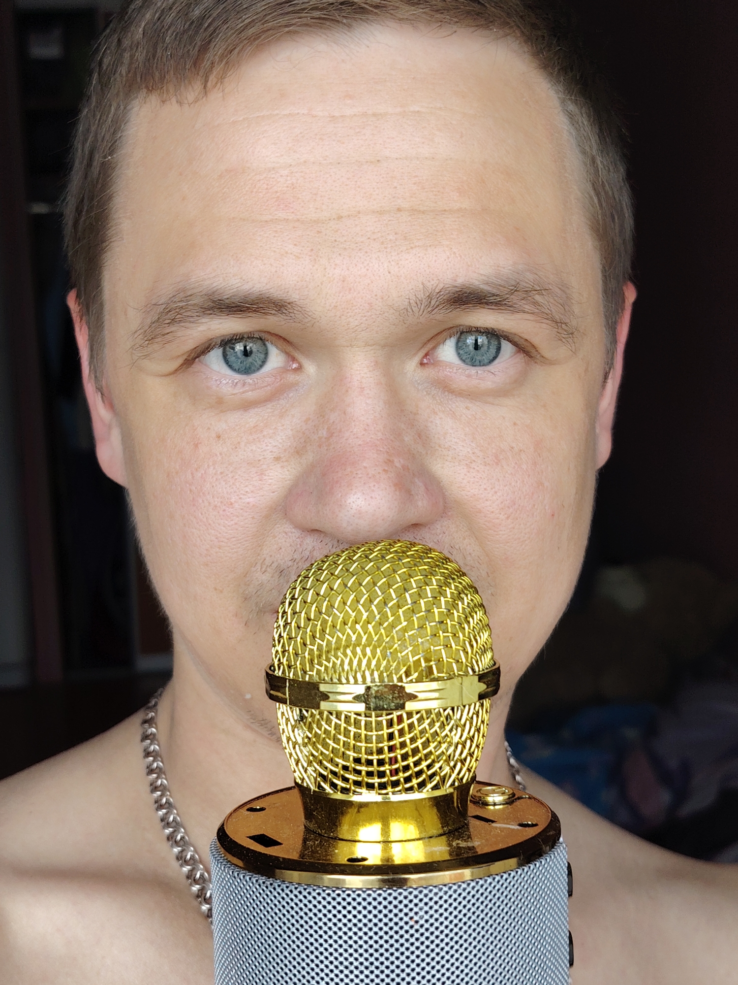 A close-up, chest-up portrait features a light-skinned individual, likely male, looking directly at the camera with clear blue eyes. The person has short brown hair, visible forehead wrinkles, and subtle stubble around the mouth and chin. A prominent gold-colored microphone with a mesh head is positioned centrally, almost touching the person's mouth. The microphone's base is silver-grey with a textured, perforated surface. A silver-colored chain necklace is visible around the individual's neck, resting against bare skin. The background is dark and out of focus, suggesting an indoor setting. The individual's expression is neutral and direct. The image was captured in Chaikovsky, Russia.
