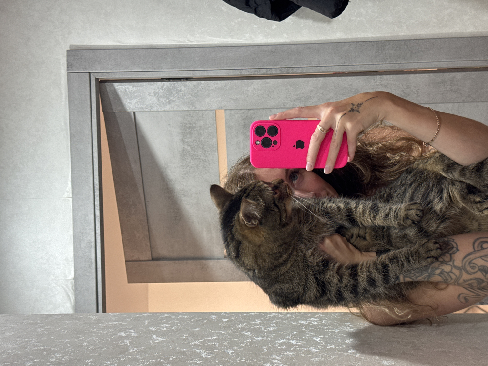 A person with long, curly brown hair holds a brown tabby cat close to their face while taking a selfie in a mirror. The person is holding a bright pink iPhone. The person has a bracelet on their right wrist, a ring on their right hand, and tattoos on both arms. The background is a gray wall and a countertop with a speckled pattern. A black jacket hangs in the top portion of the mirror's reflection. The image was likely taken indoors. It is not possible to determine the location of this image as Chaikovsky, Russia, without additional information.