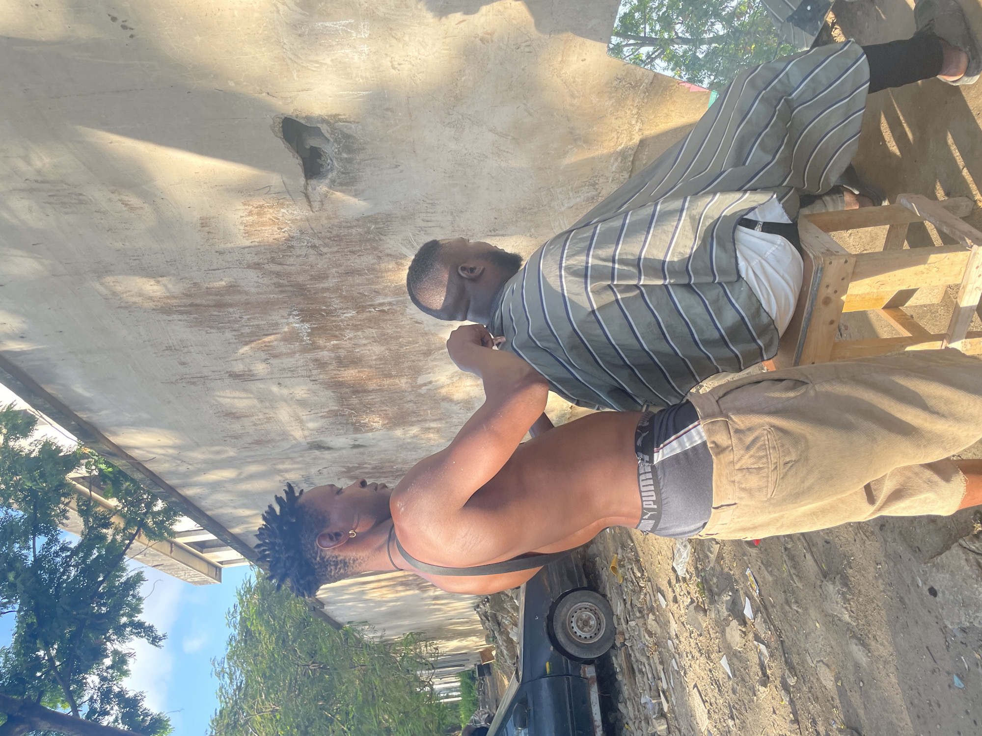 Two men are outdoors in Ikoyi, Nigeria, near a weathered building. One man, shirtless with khaki pants, stands with his back to the camera, looking at another man sitting on a wooden stool. The seated man wears a striped shirt. A car is visible in the foreground. The environment consists of trees, blue sky, and ground with debris.