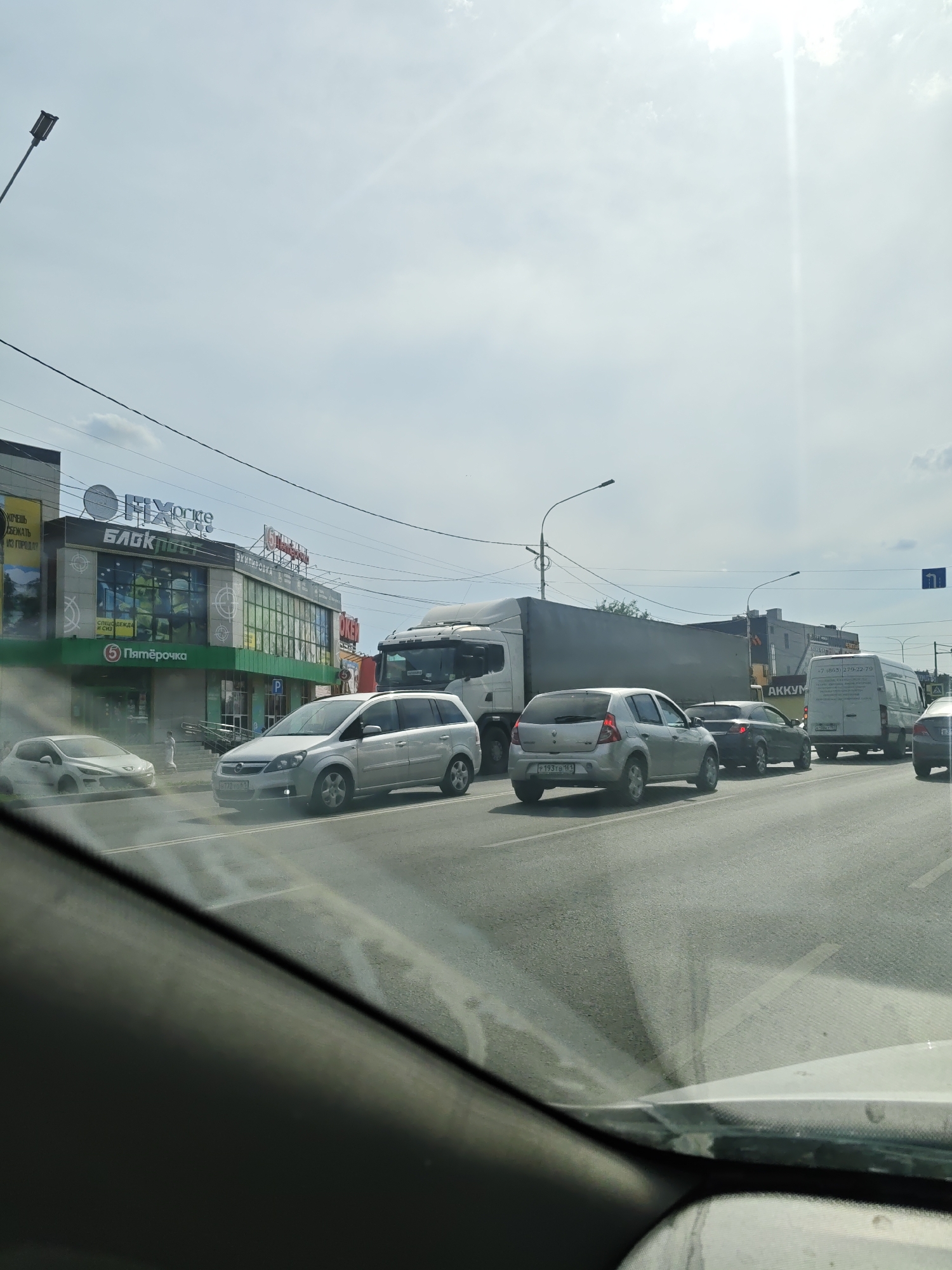 The image depicts a street scene in Novocherkassk, Russia, likely on a sunny day with a partly cloudy sky. The foreground shows the interior of a vehicle, with the dashboard and windshield visible. In the middle ground, several cars are on a multi-lane road, some of which appear to be in traffic. On the right side of the road, there is a white van with text on its side, and further down the road, another vehicle. On the left side of the road, there is a building complex with several storefronts. One store has a prominent green awning and signage that reads "Пятёрочка" (Pyaterochka). Another store has signage that includes "FIX PRICE" and "БЛОКПОСТ" (Blockpost). There are also streetlights and power lines visible in the image. The overall impression is of a typical urban street with commercial activity and vehicular traffic.