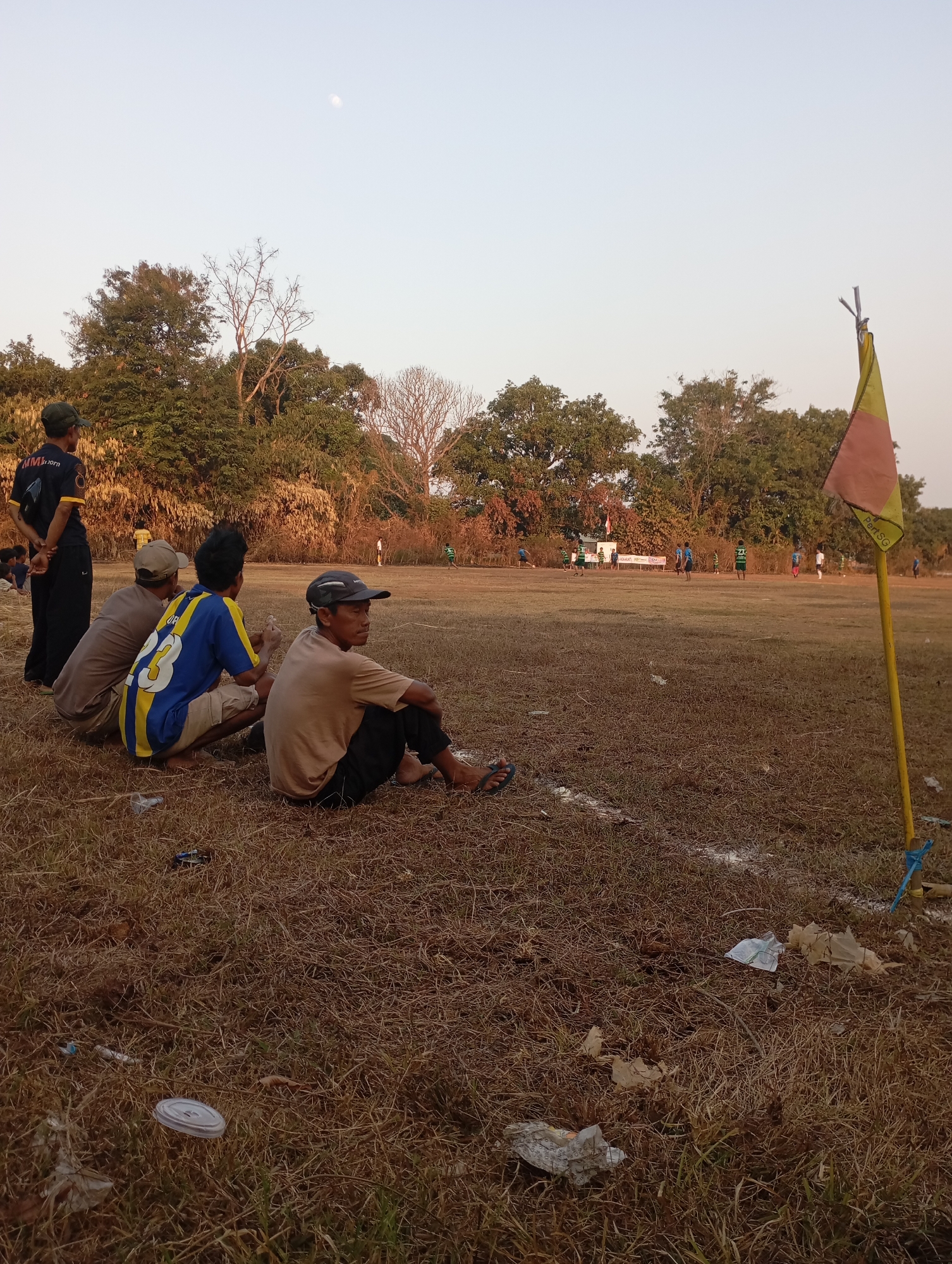In the late afternoon light of Kecamatan Haurgeulis, Indonesia, a group of four men are seated on dry, matted grass, facing a sparsely populated soccer field. The men are casually dressed, some with hats, observing a soccer game in progress in the distance. A corner flag with a yellow and red banner stands to the right of the frame, planted in the ground with blue ties. The field itself appears worn, with patches of dry earth and sparse grass, marked by a white line. Trees and foliage form a backdrop on the left side of the image, and more trees are visible further down the field. The sky is a pale, clear blue with a faint crescent moon visible. Scattered litter, including plastic cups and wrappers, lies on the grass in the foreground.