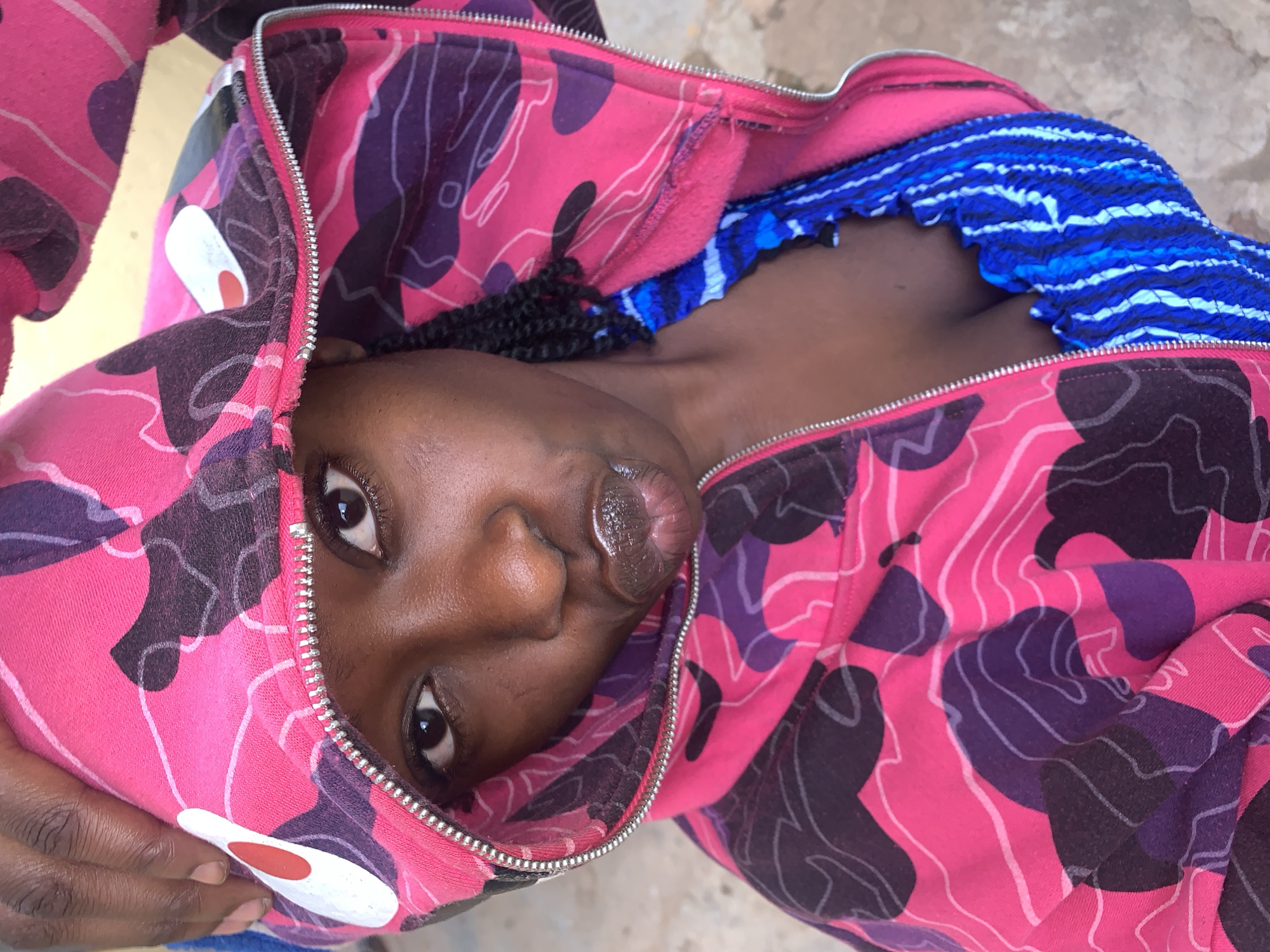 An overhead, close-up photograph depicts a young individual with dark skin, positioned in what is described as Rafin Par, Nigeria. The subject looks directly into the camera with dark eyes and is making a distinct pout with glossy lips. They wear a full-zip hoodie featuring a prominent pink, purple, and black camouflage pattern, characteristic of the BAPE brand, with a visible shark-face design element (eye and mouth area) on the left side of the hood. The hoodie's silver-toned zipper is partially visible, extending from the bottom of the frame upwards. The individual's right hand, with fingers visible, holds the edge of the hood near the top-left of the frame, pulling it slightly forward. Dark braided hair is visible emerging from beneath the hood on the right side. Underneath the open hoodie, a blue and light blue patterned or striped top with a textured, possibly smocked, fabric is discernible across the chest. The background is out of focus, suggesting an outdoor setting with a light, muted ground surface visible. Natural lighting illuminates the scene.