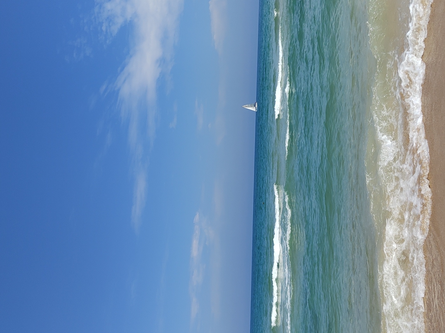 The image depicts a beach scene, likely in Tel-Aviv, Israel, based on the coastal characteristics. A bright blue sky with scattered clouds fills the left half of the frame. The right half shows the ocean, with white waves rolling towards a sandy beach. A small sailboat is visible on the horizon. There are no people visible.