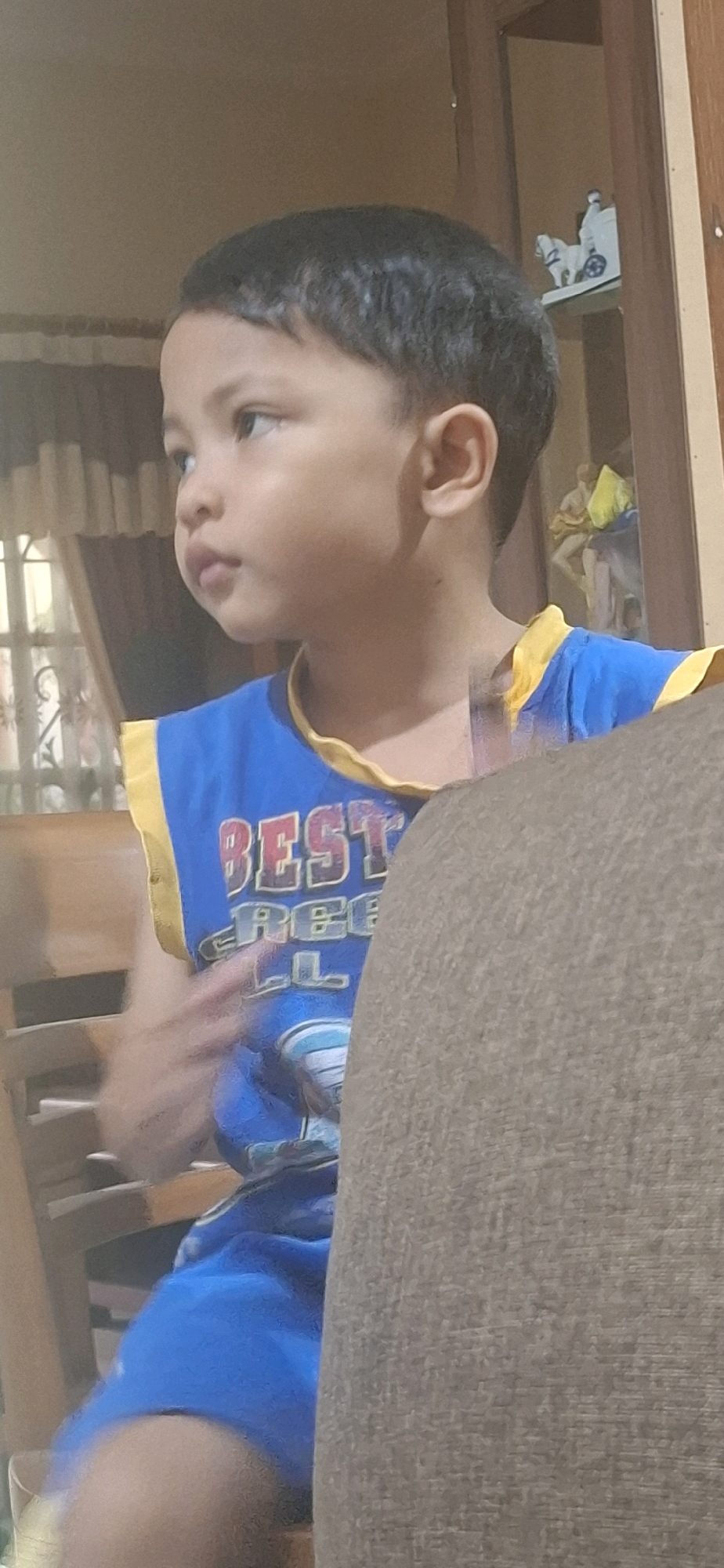 A young boy, appearing to be of East Asian descent, is shown from the chest up, looking to the left. He has dark hair and is wearing a sleeveless blue shirt with yellow trim. The shirt has text that reads "BEST" in red letters, with "FREE" and "ALL" below it in a lighter color. He is positioned in front of a brown textured surface, which appears to be the back of a sofa or chair. In the background, a wooden cabinet with glass doors is visible. Inside the cabinet, several decorative items are present, including what looks like a white horse-drawn carriage on a shelf. To the left of the boy, a window with light-colored curtains is partially visible, and wooden furniture that may be chairs is also in the frame. The lighting suggests an indoor setting, possibly during the day or evening. The image is slightly blurry, particularly the boy's arm which is in motion. The setting appears to be a domestic interior in Desa Gagaksipat, Indonesia.