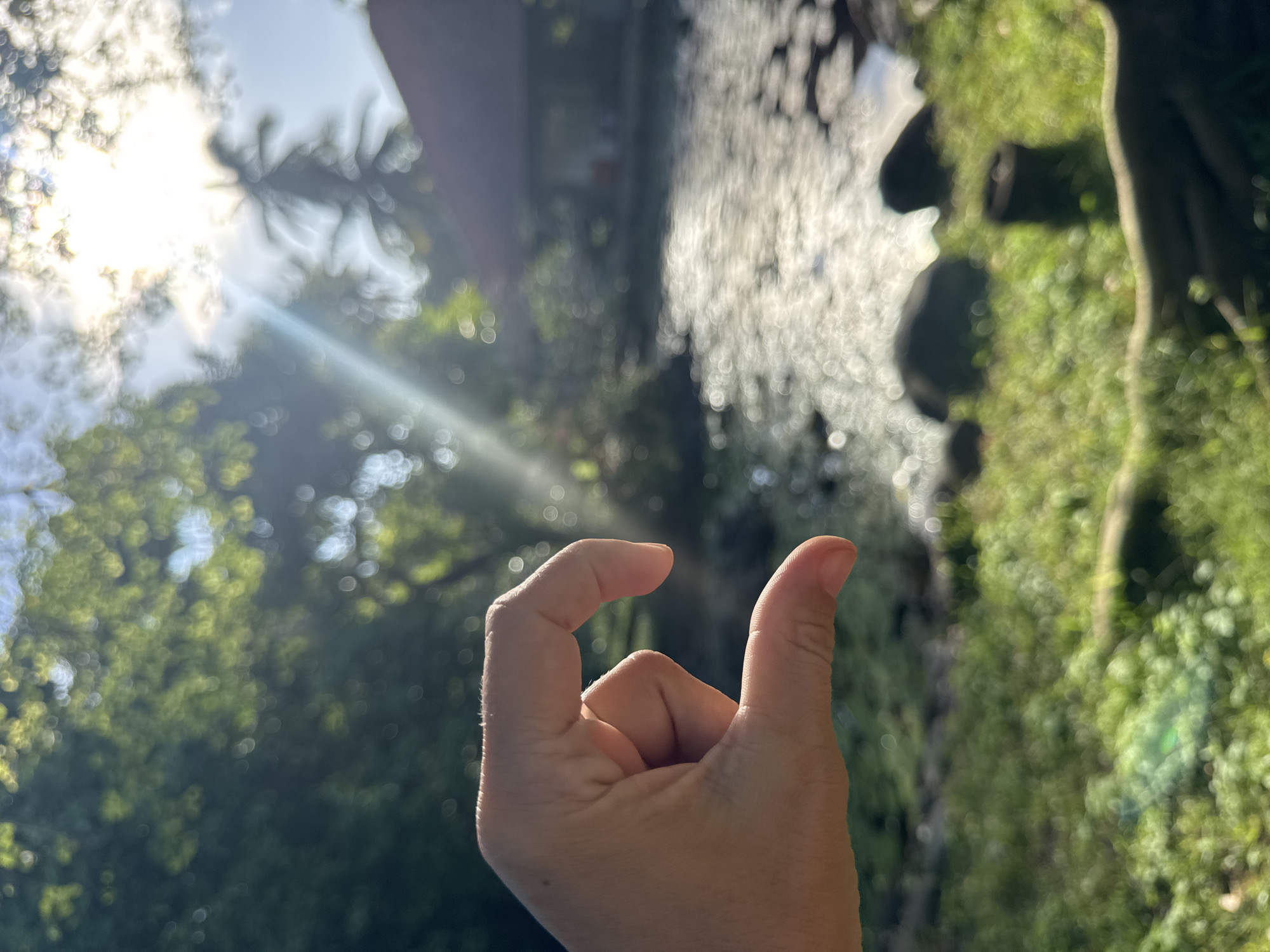 A photograph taken in Kuta Selatan, Indonesia, shows a person's hand in the foreground making a "pinching" gesture against a blurry background. The background consists of green vegetation, possibly trees, a sunlit water body (likely a stream or river), and a hint of a structure. The sunlight creates bokeh in the image, adding to the blurred effect.