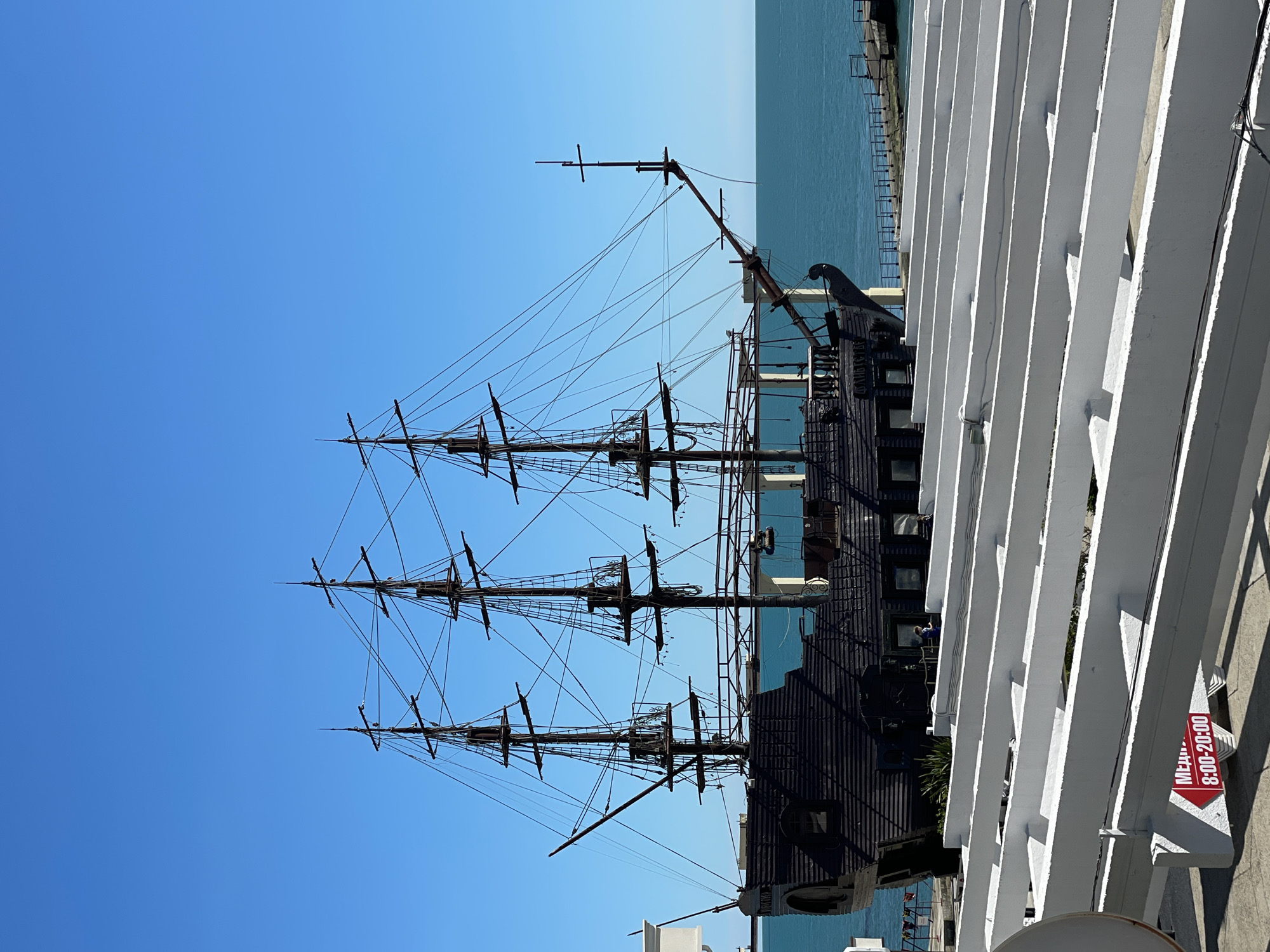 This image, taken in Sochi, Russia, showcases a large, dark-hulled sailing ship, likely a historical replica or a pirate-themed vessel, moored at a waterfront on a clear, sunny day. The ship features three tall masts with extensive rigging and cross spars, rising prominently against a bright, cloudless blue sky. Its hull is dark, contrasting with the vibrant blue of the sea stretching horizontally behind it. The calm, light blue water suggests peaceful conditions.
In the foreground, a distinct white architectural structure, possibly part of a pier, building, or pergola, frames the right side of the scene. This structure is composed of parallel white beams casting strong, defined shadows, indicating bright daylight, likely late morning or early afternoon. A small red sign is attached to one of these white beams, displaying the word "МЕНЮ" (MENU) and the operating hours "8:00-20:00", suggesting a nearby commercial establishment, perhaps a restaurant or cafe, associated with the waterfront or the ship itself.
A distant pier or breakwater is visible extending into the sea. The overall setting is a picturesque coastal environment, characteristic of a marina or port designed for tourism. There are no visible people, but the sign implies active commerce. The image captures a serene moment with the historic-looking ship as the focal point against a vivid blue backdrop.