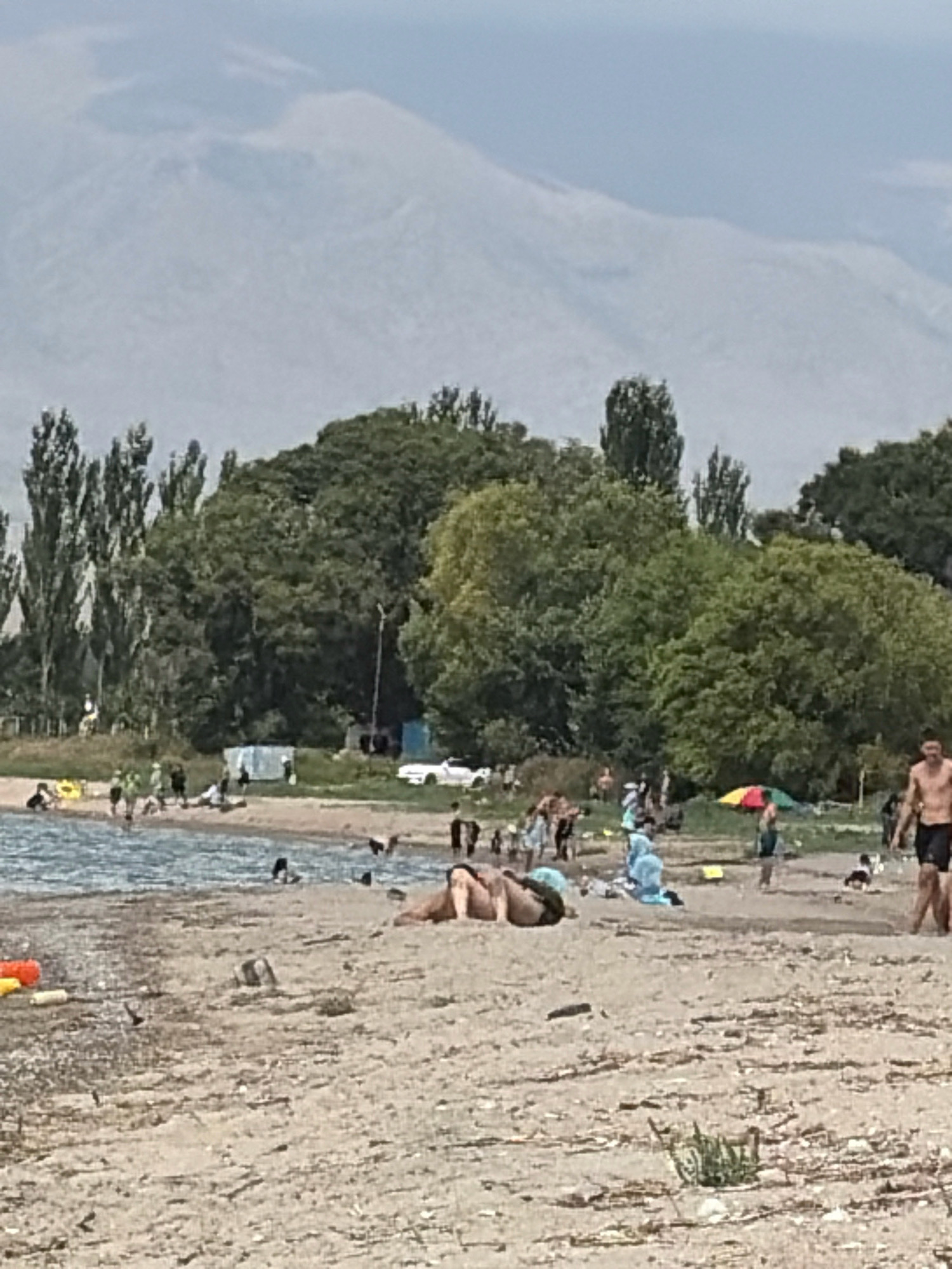 A wide shot depicts a sandy beach on a cloudy day. The background shows a hazy mountain range, with a treeline and some buildings separating it from the beach. The foreground features a body of water with several people swimming and wading. On the beach, there are many individuals scattered around, some sunbathing, some standing and conversing, and some walking. Objects like beach toys and umbrellas are also visible. The overall atmosphere suggests a recreational outing at a beach resort.
