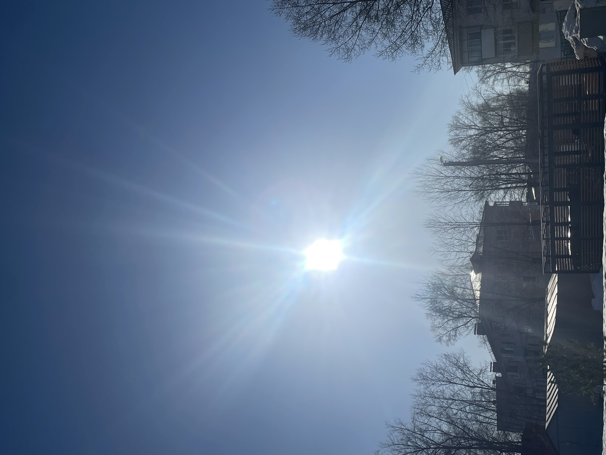 The image shows a bright, sunny winter day in Chaikovsky, Russia. The sun is high in the sky, creating intense glare. Buildings and bare trees line the right side of the frame, covered in snow. A fence is visible in the foreground, also partially covered with snow. The left side of the image shows a clear blue sky. No people are visible.