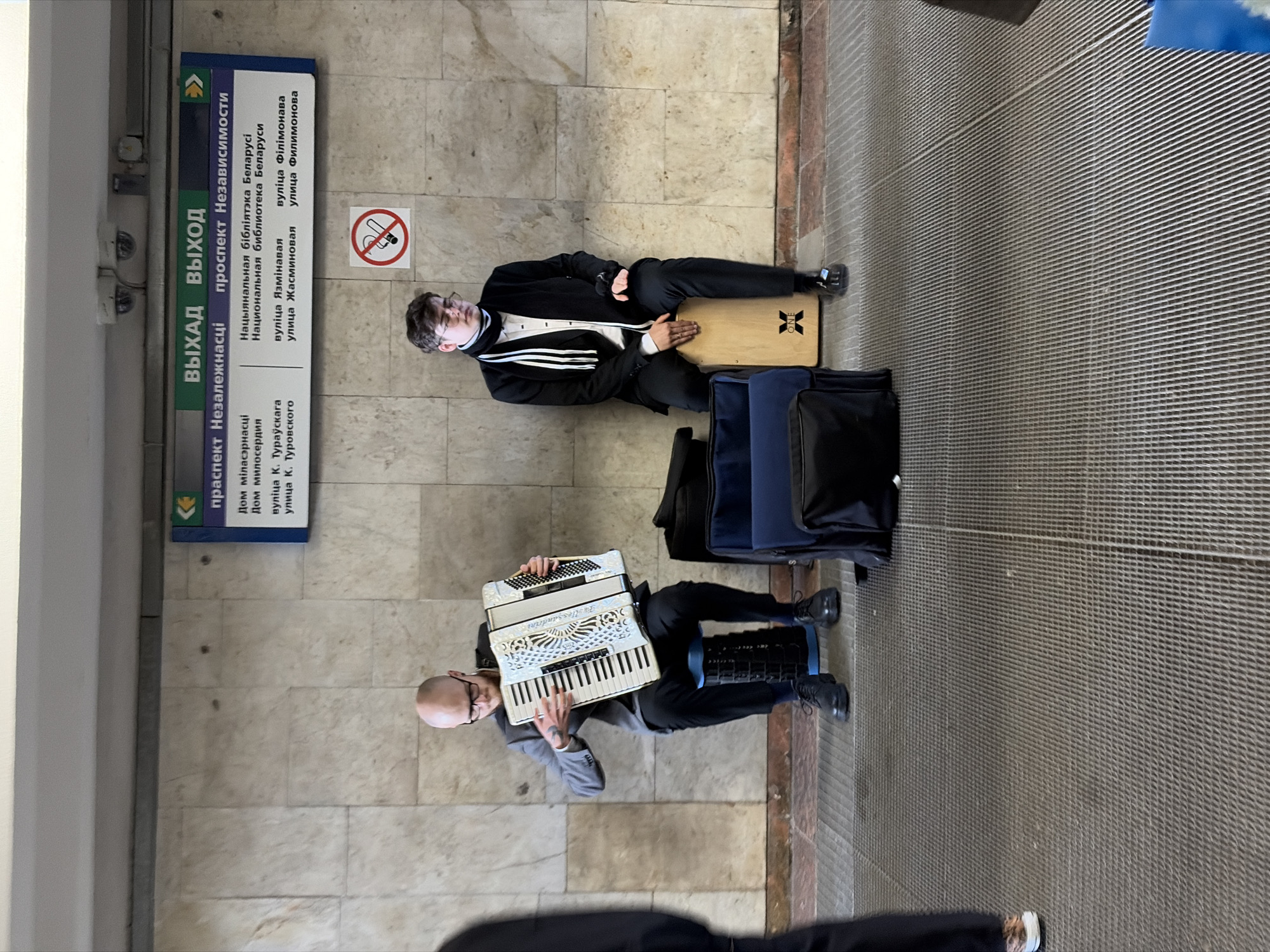 Here is a forensic description of the image:
The image depicts two buskers performing on the floor of what appears to be a subway or train station. The man closest to the camera is playing a white accordion. He is bald, wearing glasses, a gray blazer, and black pants with black shoes. He has tattoos on his right arm. To the side, there is another man sitting on a wooden cajon. He is wearing a black suit, a white shirt, and a black and white scarf. Both men have gear, including bags, next to them. A sign behind them reads "выхад", "выход", and "independence avenue" in both Belarusian and Russian. There is also a no smoking sign. The location appears to be a public transit area in an unknown city.