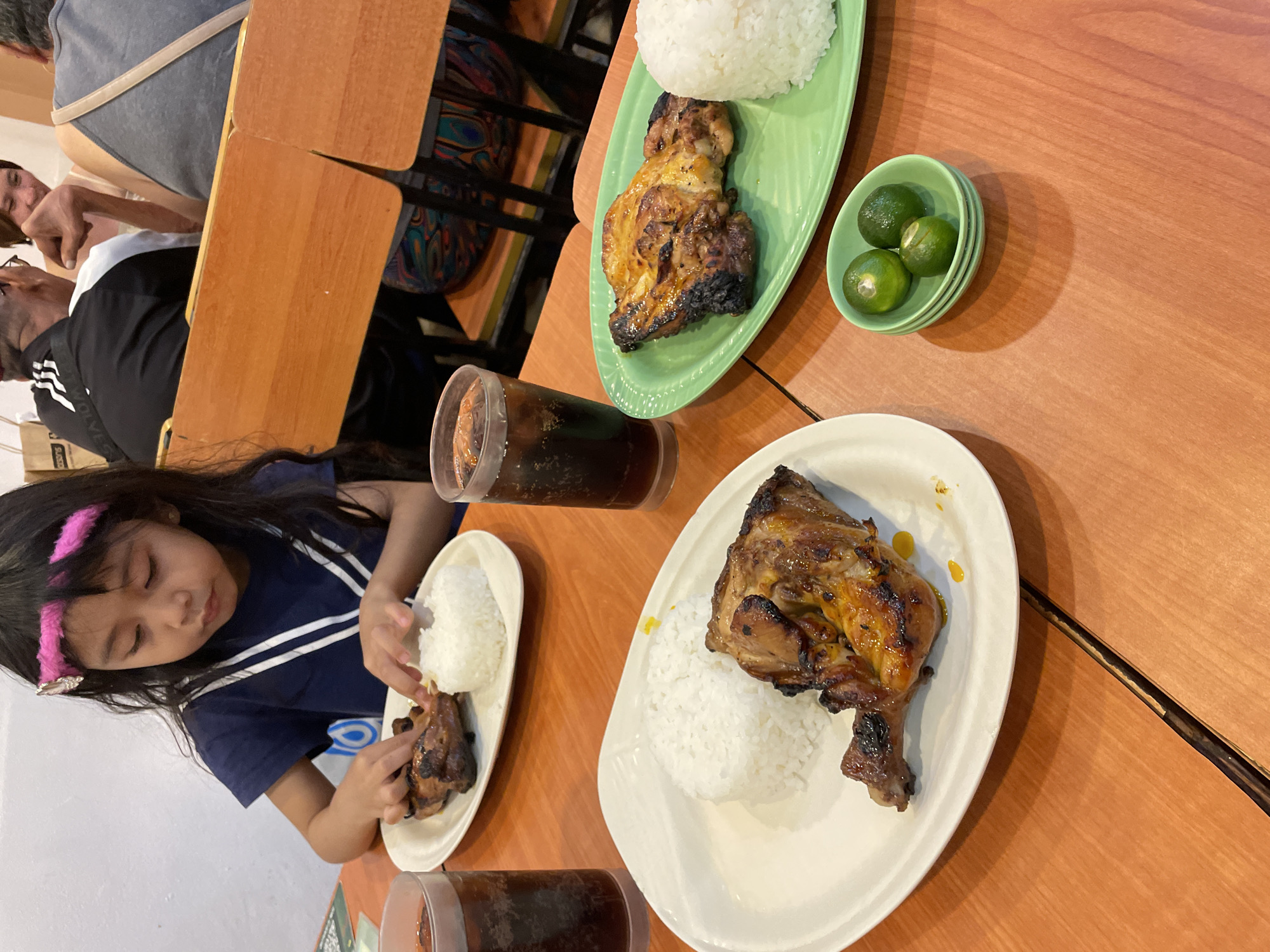 The image depicts an interior scene inside a casual dining restaurant, identified as a Mang Inasal branch in Davao City, Philippines. In the foreground, a young Asian girl with long dark hair, adorned with a pink furry headband, is seated at an orange wooden table. She wears a navy blue top with white stripes and is actively engaged in eating, using her hands to pull apart a piece of grilled chicken from a white oval plate, which also contains a mound of white rice. A tall glass of dark soda with ice is positioned to her left.
To the right of the girl's plate, another identical white oval plate with grilled chicken and rice is visible. Further right, in the foreground, a green oval plate holds another serving of grilled chicken and white rice, alongside a second tall glass of dark soda. A small green dish containing three small, whole green citrus fruits, specifically calamansi, sits on the table, serving as a condiment.
In the background, several adults are seated at similar orange wooden tables and chairs. A man, seen from behind, wears a black and white athletic jacket and appears to be interacting with a woman whose smiling face is partially visible. Another individual in a grey tank top is seated on the far right. The restaurant features light-colored walls, and a blue, circular logo is faintly visible on the wall behind the girl.