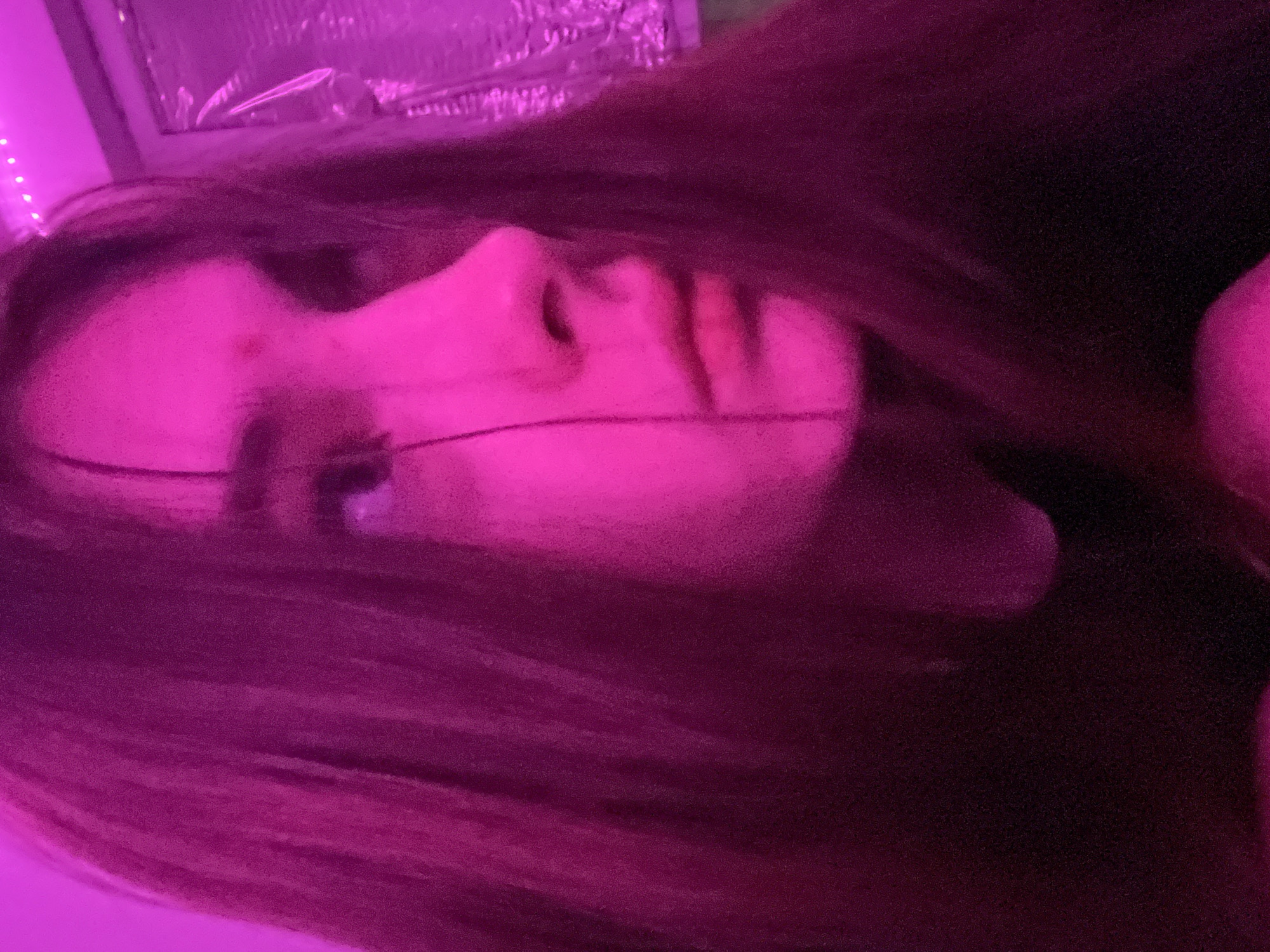 A close-up, indoor shot features a person with long brown hair, bathed in pinkish-purple light. The person is lying down, with their face turned towards the side. The scene also includes part of a window and possibly LED string lights. No evidence is present to indicate the location of the photograph is in Saint Petersburg, Russia.