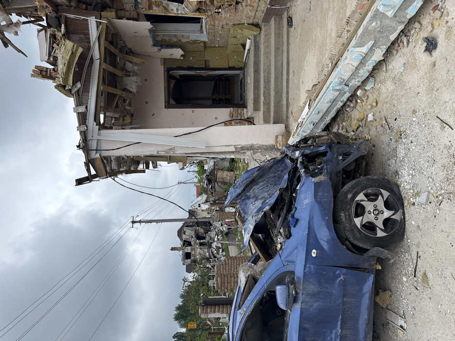The scene depicts a war-torn area, with a severely damaged blue car in the foreground. It is positioned next to a building that has suffered significant destruction, exposing its inner structure and insulation. In the background, further wreckage and debris are visible, suggesting widespread devastation. The sky is overcast, adding to the somber and bleak atmosphere.