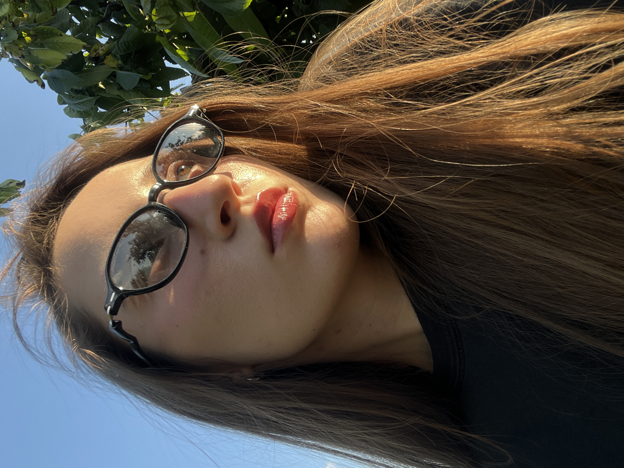 A woman with long, brown hair and black-rimmed glasses is shown in a close-up, outdoor portrait. She is looking to the side, with sunlight illuminating her face and highlighting her lips. The background shows a clear blue sky and green foliage from a tree. The woman is wearing a black shirt. The setting appears to be a park or garden, possibly during late afternoon or early evening due to the warm lighting.