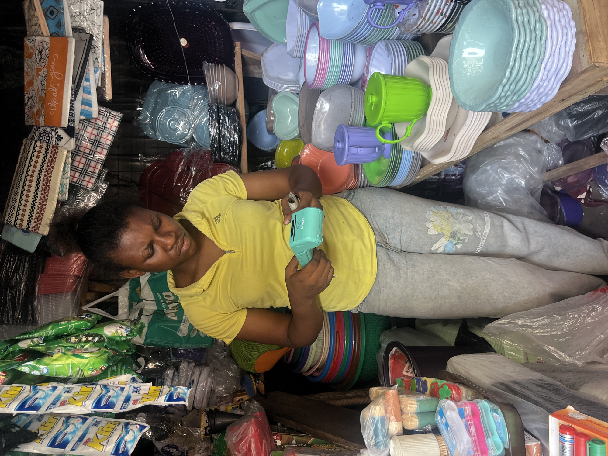 This image captures a busy market stall or small shop in Nkpor, Nigeria, filled with a dense array of household goods. The central subject is a young woman, likely the vendor, seen from the waist up. She wears a yellow short-sleeved top with an "adidas" logo and light grey sweatpants adorned with a faded floral pattern. Her expression is one of focus as she holds a small, light-blue device in her hands, possibly a mobile payment terminal or a cash-counting machine, with the word "Opay" clearly visible on its side.
The scene is dominated by the merchandise, which is tightly packed around her and stacked on shelves. A prominent feature is the large quantity of colorful plastic kitchenware, including bowls, plates, and cups in shades of blue, green, pink, purple, and white. There are also plastic baskets, serving trays, and various other containers.
Among the other visible products are numerous packets of detergent. Text on these packages includes "Klin," "CONCENTRATED DETERGENT 2X," and phrases like "dry & soft." There are also stacks of paper products or bags labeled "Thank yous," bundled toothpicks, plastic cutlery, and other general merchandise, much of it wrapped in clear plastic. The cluttered but organized nature of the stall suggests a well-stocked local business. The lighting indicates an indoor setting or a covered outdoor space typical of market environments.