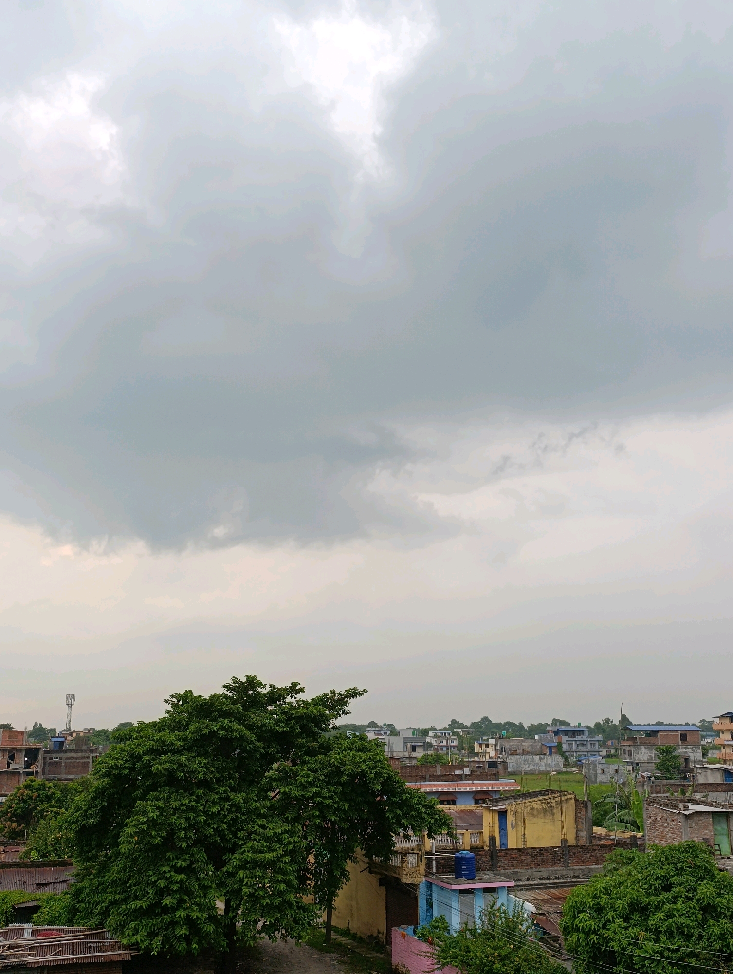 The image depicts an overcast sky over a densely populated urban area, likely a neighborhood in Biratnagar, Nepal. The sky is filled with heavy, gray clouds, suggesting impending rain. Below the clouds, a cluster of buildings with varied architectural styles, predominantly concrete and brick structures, are visible. These buildings appear to be residential and commercial, with some showing signs of wear and tear. Green trees, some large and leafy, are interspersed among the buildings, adding a touch of nature to the urban landscape. A telecommunications tower can be seen in the distance. The overall scene conveys a sense of everyday life in a tropical or subtropical urban environment under a cloudy sky.