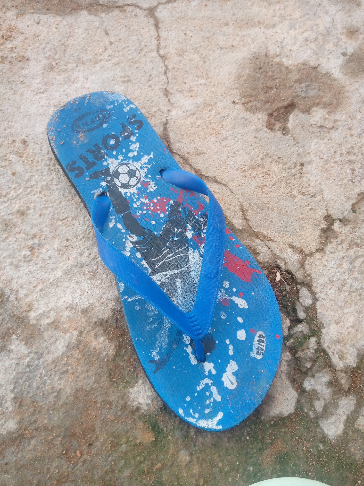 A blue flip-flop with a soccer-themed graphic lies on a cracked concrete surface. The graphic on the flip-flop depicts a soccer ball, a silhouette of a soccer player, and abstract paint splatters in red and white. The flip-flop appears worn and has some dirt on it. The concrete surface shows signs of age and weathering, with visible cracks and discoloration. The overall scene suggests an outdoor setting, possibly a street or courtyard, in Jalingo, Nigeria.