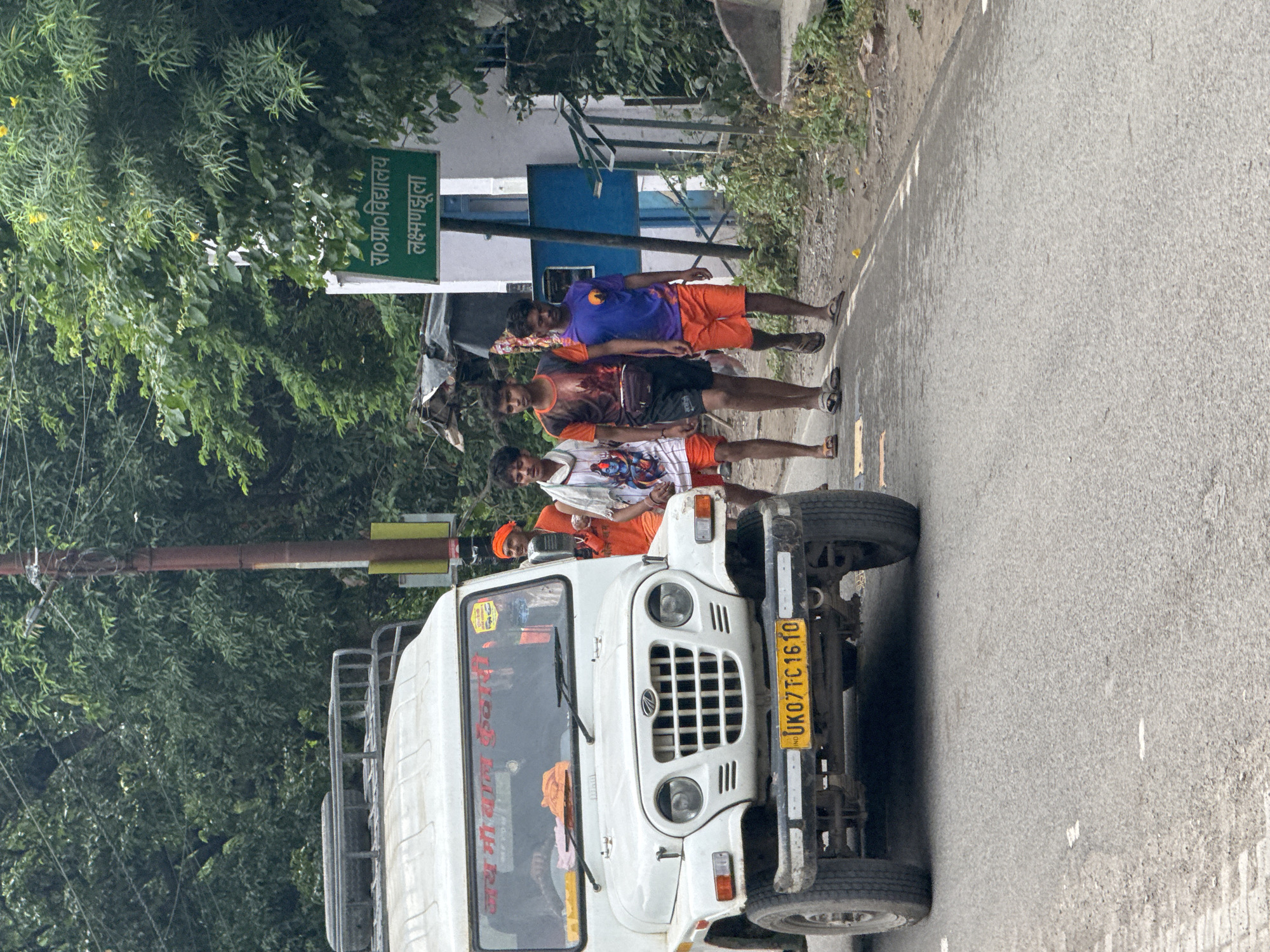 The image shows a white Mahindra Thar jeep parked on a road in Rishikesh, India. The jeep has a roof rack and a yellow license plate that reads "UK07TC1610". There are several people walking on the road next to the jeep. The people appear to be young men. One person is wearing an orange turban and appears to be a sadhu. There is a green sign in the background with Hindi text. The environment appears to be a roadside with trees and vegetation.