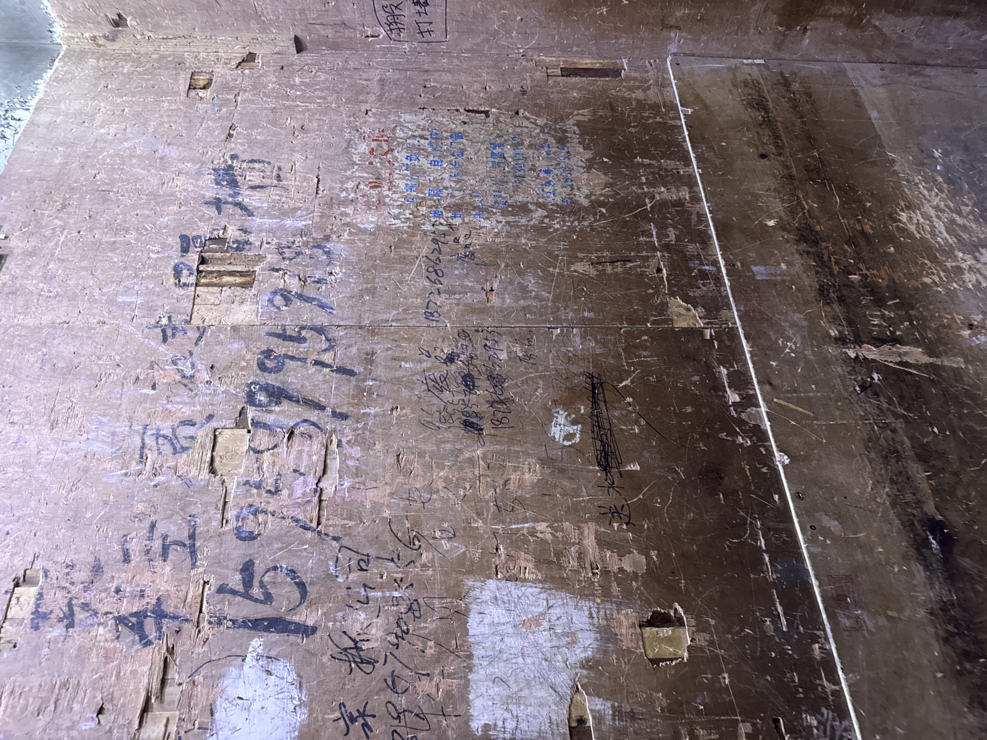 The image captures a close-up view of a weathered, wooden wall with graffiti and handwritten text. The wall appears to be made of plywood or similar material, showing signs of age and wear, including visible grain, splintering, and some small holes. The graffiti is in Chinese and includes numbers and characters.
In the upper left quadrant, large, bold black characters are visible, possibly indicating a business or location. Below this, a series of large numbers "15 23 39 39" are scrawled in black. To the left of these numbers, there are more handwritten Chinese characters.
In the center and right portions of the image, the text becomes more dense and varied in color, with some blue and black ink. Some of the text appears to be contact information or advertisements, with phone numbers like "13726862911" visible. Red ink also appears in a circular or oval-shaped area in the upper right quadrant. In the far right, a black box with Chinese characters is present, likely a stamp or sign.
The lower portion of the image shows a horizontal element, possibly a shelf or a piece of furniture, made of a darker material with a reflective surface, partially obscuring the lower part of the wall. There are also horizontal lines and scratches across the wooden surface, suggesting it is an interior wall in a somewhat dilapidated or functional space.
The scene is consistent with typical urban graffiti found in areas like Yiwu, China, known for its bustling trade markets and associated signage and markings. The wall likely serves as a bulletin board or advertising space within a commercial or storage area.