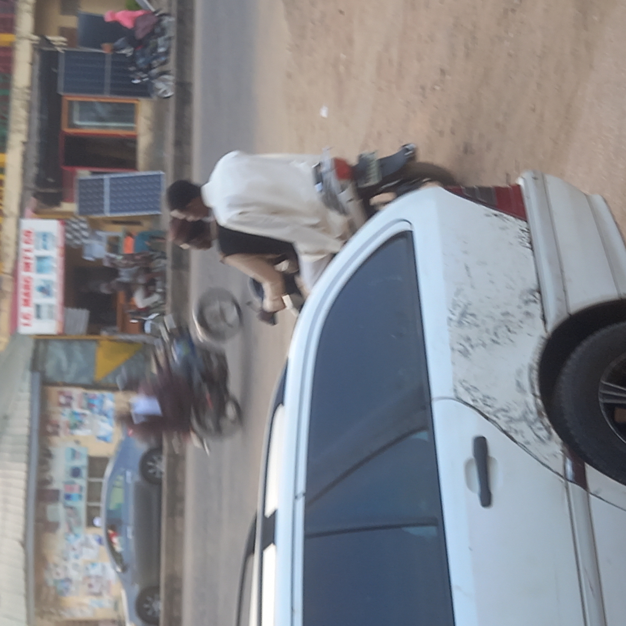 The image shows an outdoor street scene in Dadi Riba, Nigeria. A white car is parked on the side of the road. A motorcycle with two people on it drives past the car. Buildings line the street on the left. One building has solar panels.