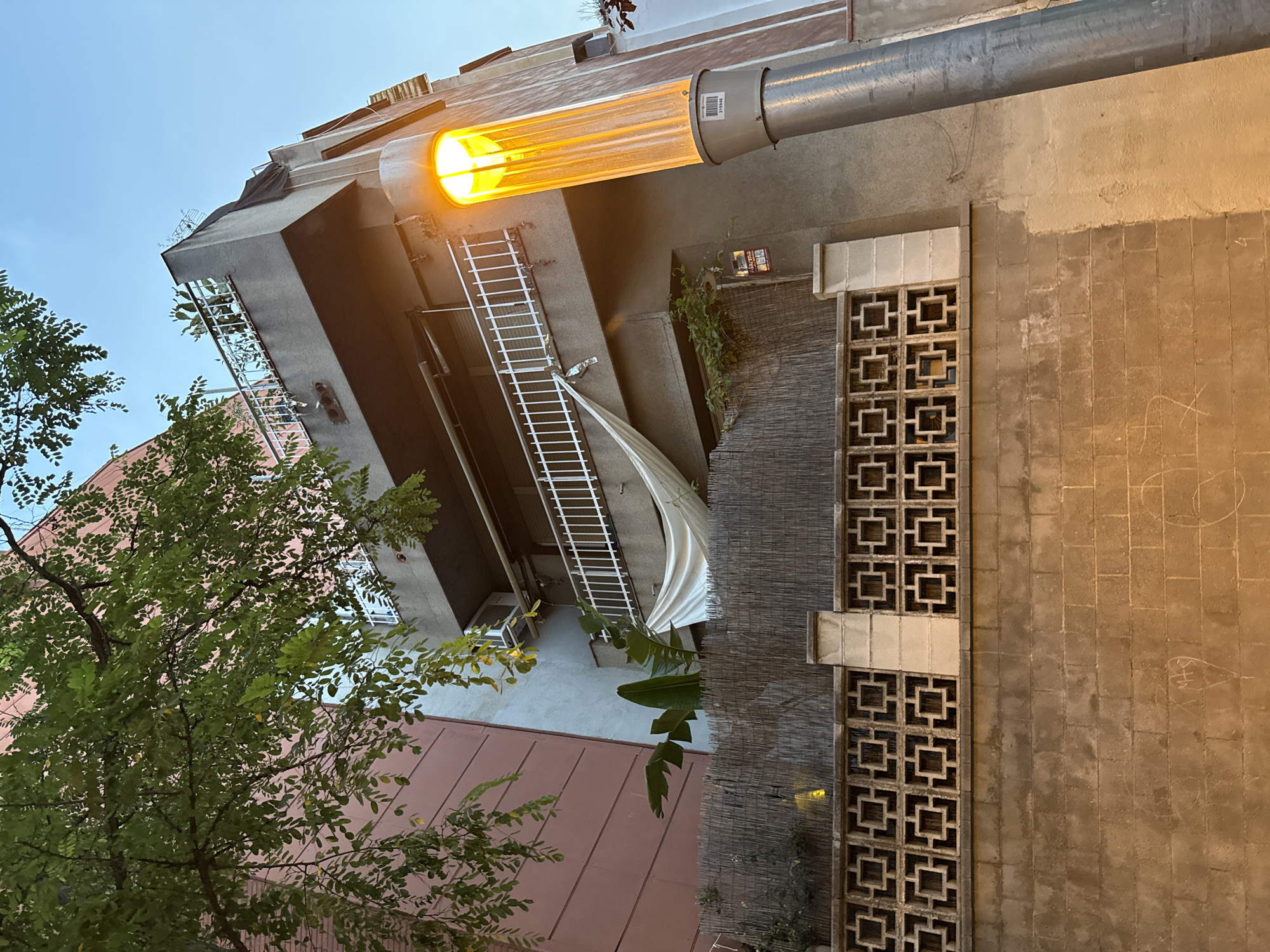 The image depicts a street scene in Barcelona, Spain, at dusk. In the foreground, a rough-textured concrete wall is visible, with faint chalk markings. Above this wall is a decorative concrete block fence with a geometric pattern. Behind the fence is a bamboo screen. A modern building with several balconies rises in the background. The building has a reddish-pink facade on the left and a gray facade on the right, with a prominent balcony on the top floor adorned with plants. A tall, cylindrical street lamp with a bright orange light is positioned in the right foreground, casting a warm glow. A white sheet is draped over a railing on one of the balconies, and an air conditioning unit is visible on the side of the building. Lush green foliage from a tree hangs down from the upper left of the frame. The sky above is a clear, pale blue, indicating the transition from day to night.