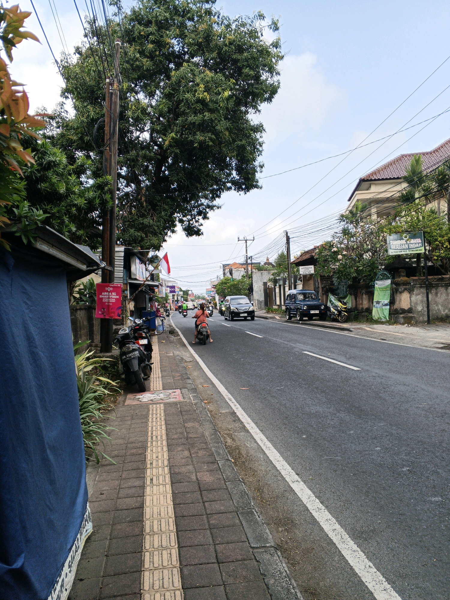 The image depicts a street scene in Denpasar, Indonesia, likely during daylight. A two-lane asphalt road runs diagonally through the frame, bordered by a pedestrian sidewalk on the left. The sidewalk is paved with rectangular tiles, featuring a tactile warning strip. On the left side of the sidewalk, a blue tarp is draped over a structure, partially obscuring a concrete wall and some greenery. A utility pole with numerous wires stands prominently near the sidewalk.
Several parked motorcycles are visible along the sidewalk. Further down the street, traffic moves in both directions. On the road, a white sedan, a dark-colored SUV, and multiple motorcycles are in motion. A person is riding a pink motorcycle in the right lane, heading away from the viewer. Buildings line the right side of the street, some with tile roofs and balconies. Greenery, including trees and flowering plants, is abundant. Signs with Indonesian text are visible on some buildings. The sky is partly cloudy.