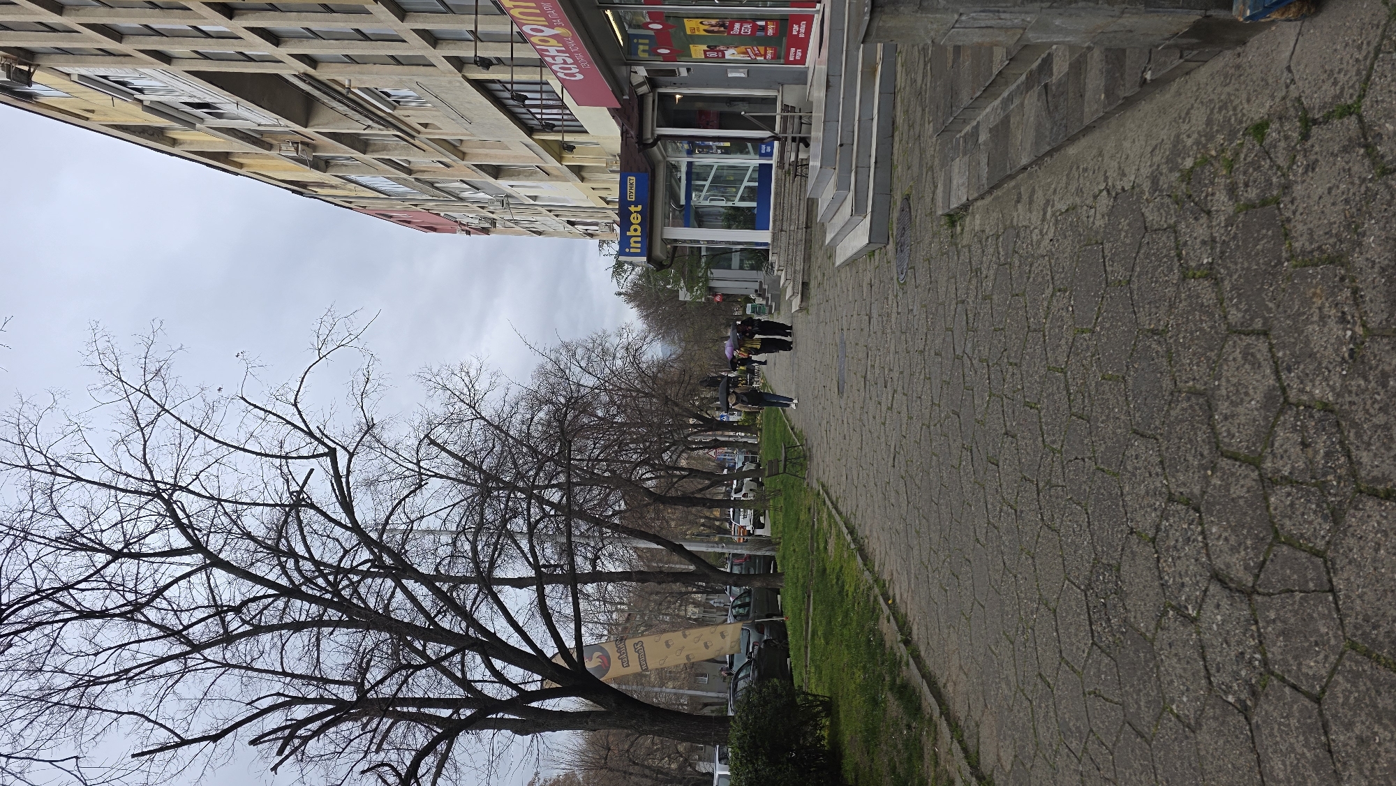 The image shows a street scene in Blagoevgrad, Bulgaria. On the right is a paved sidewalk made of hexagonal pavers with some grass growing in the cracks. Adjacent to the sidewalk is a strip of grass, then a row of bare trees. Cars are parked along the street. Several people are walking on the sidewalk in the distance, one of whom is holding an umbrella. On the right are buildings, including establishments such as "Cashpoint" and "Inbet". The sky is overcast.