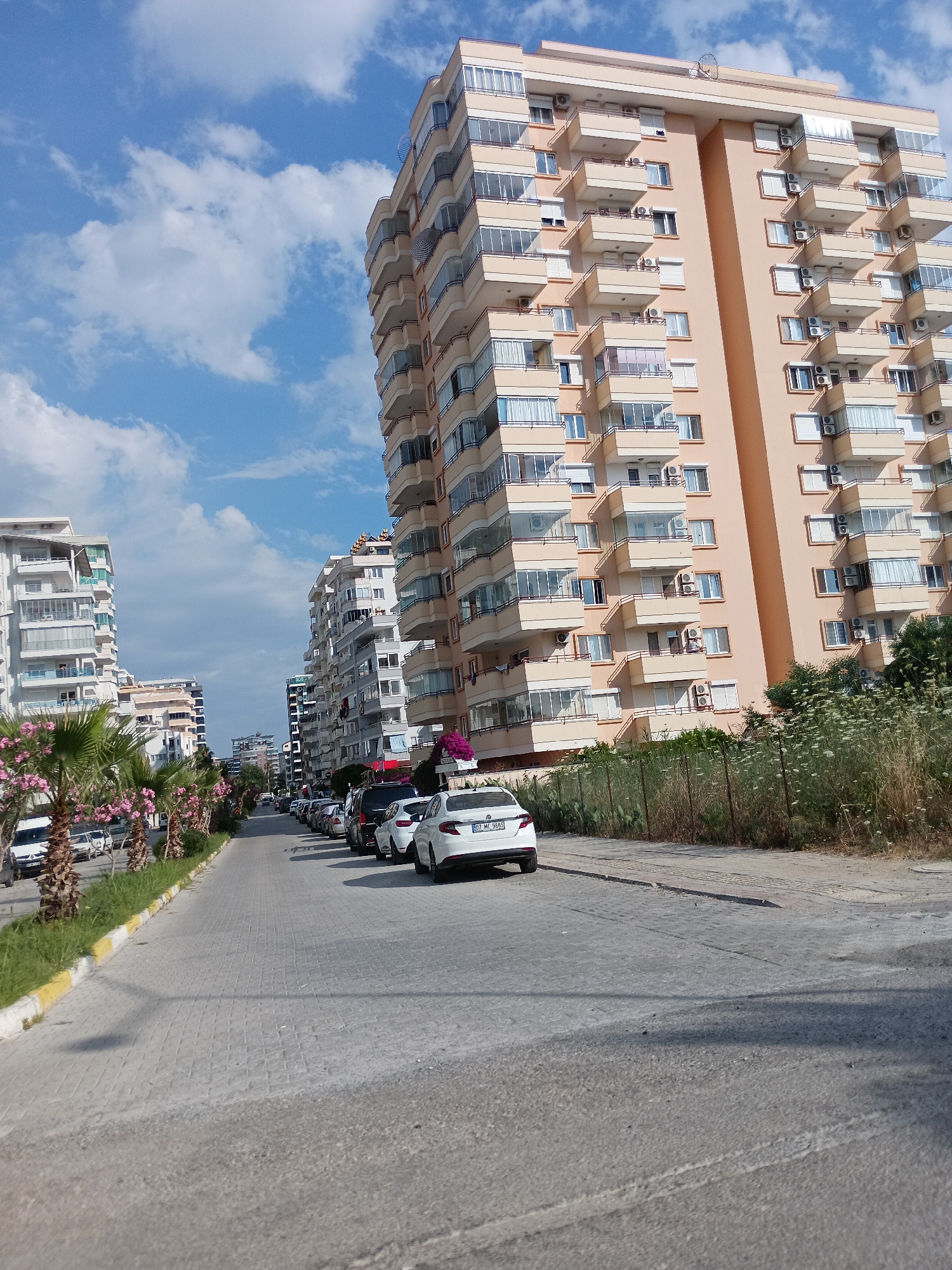 This media file captures a street scene in Alanya, Turkey, bathed in bright daylight under a partly cloudy sky. The perspective is from a street-level view, looking down a paved road lined with parked cars and tall residential buildings.
The prominent subjects are the multi-story apartment buildings. The building on the right is a tall, peach-colored structure with numerous balconies, many of which are enclosed with glass and have air conditioning units visible. The building on the left appears to be white and silver, also featuring multiple levels and balconies. Several other apartment buildings of varying heights and architectural styles are visible further down the street, creating a dense urban landscape.
Parked cars fill the right side of the street, including several white vehicles and darker SUVs. One white car in the foreground has a visible license plate that reads "07 MC 3685". Palm trees with pink flowers line the left side of the road, interspersed with green grass and a yellow-and-white striped curb. In the background, more buildings and trees are visible, suggesting a well-populated area.
The scene appears to be one of a typical day in a residential neighborhood. There are no people visibly present in the immediate vicinity, giving the impression of a quiet moment. The bright sunlight and visible clouds suggest fair weather. The presence of palm trees and the architecture of the buildings, along with the license plate format, are strong location cues for Alanya, Turkey. The overall mood is peaceful and mundane, reflecting a normal urban environment.
