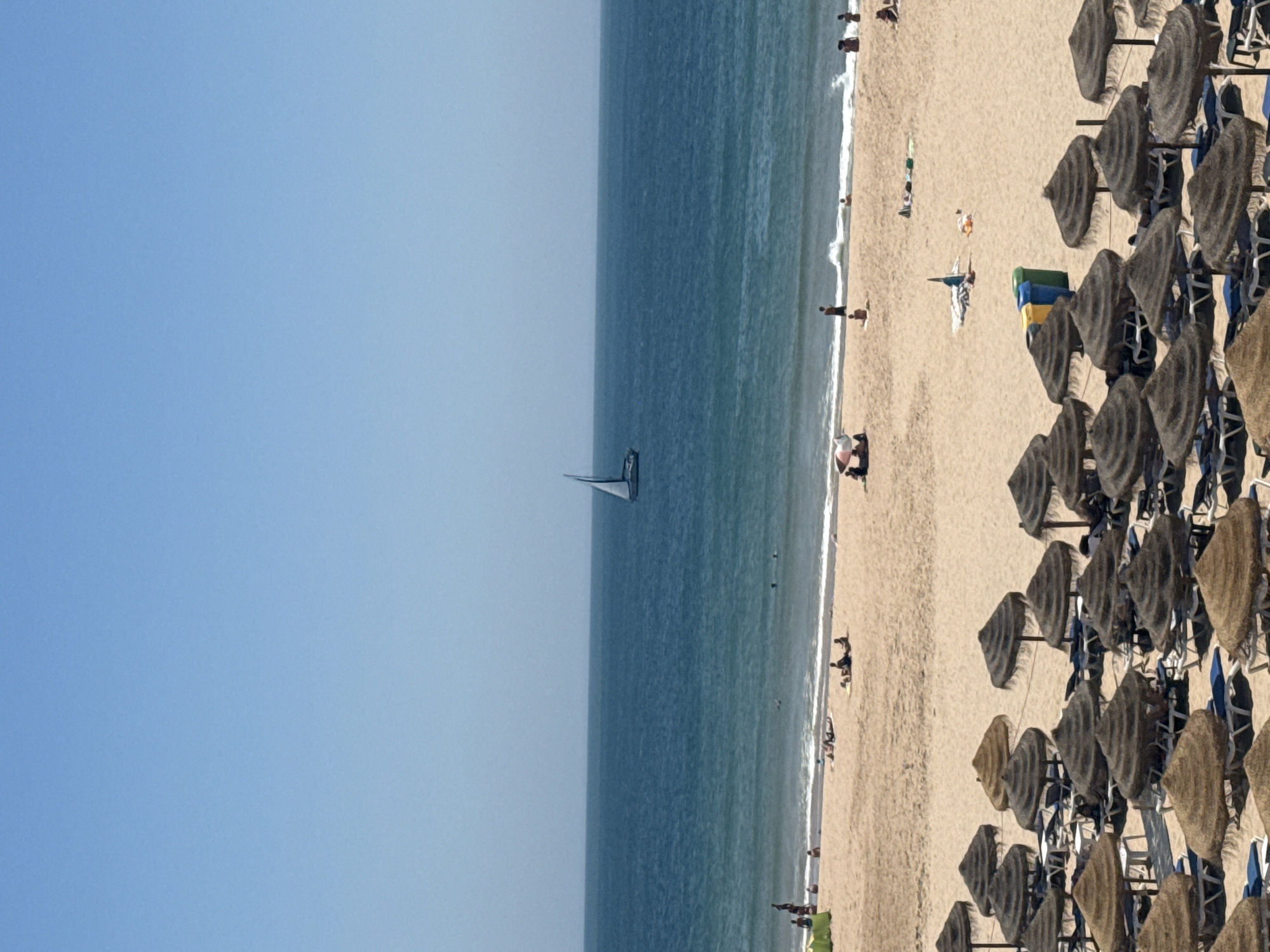This is a daytime, high-angle, full shot of a beach scene in Cascais, Portugal. The image is dominated by a large expanse of calm, blue ocean under a clear, pale blue sky. A single white sailboat with two sails is visible in the middle distance, to the left of the image center. The beach, composed of light brown sand, stretches across the foreground and middle ground. The wet sand near the waterline shows some small waves breaking.
On the beach, there are several groups of people. Some are sunbathing on towels or loungers, while others are standing or walking. Near the water's edge, a few individuals are seen interacting with the waves.
In the foreground, rows of thatched beach umbrellas with attached lounge chairs are neatly arranged. Some of the lounge chairs are occupied, while others are empty. To the right of the image, two blue trash bins and one yellow trash bin are visible among the umbrellas. The overall atmosphere is one of leisure and a warm, sunny day at the coast.