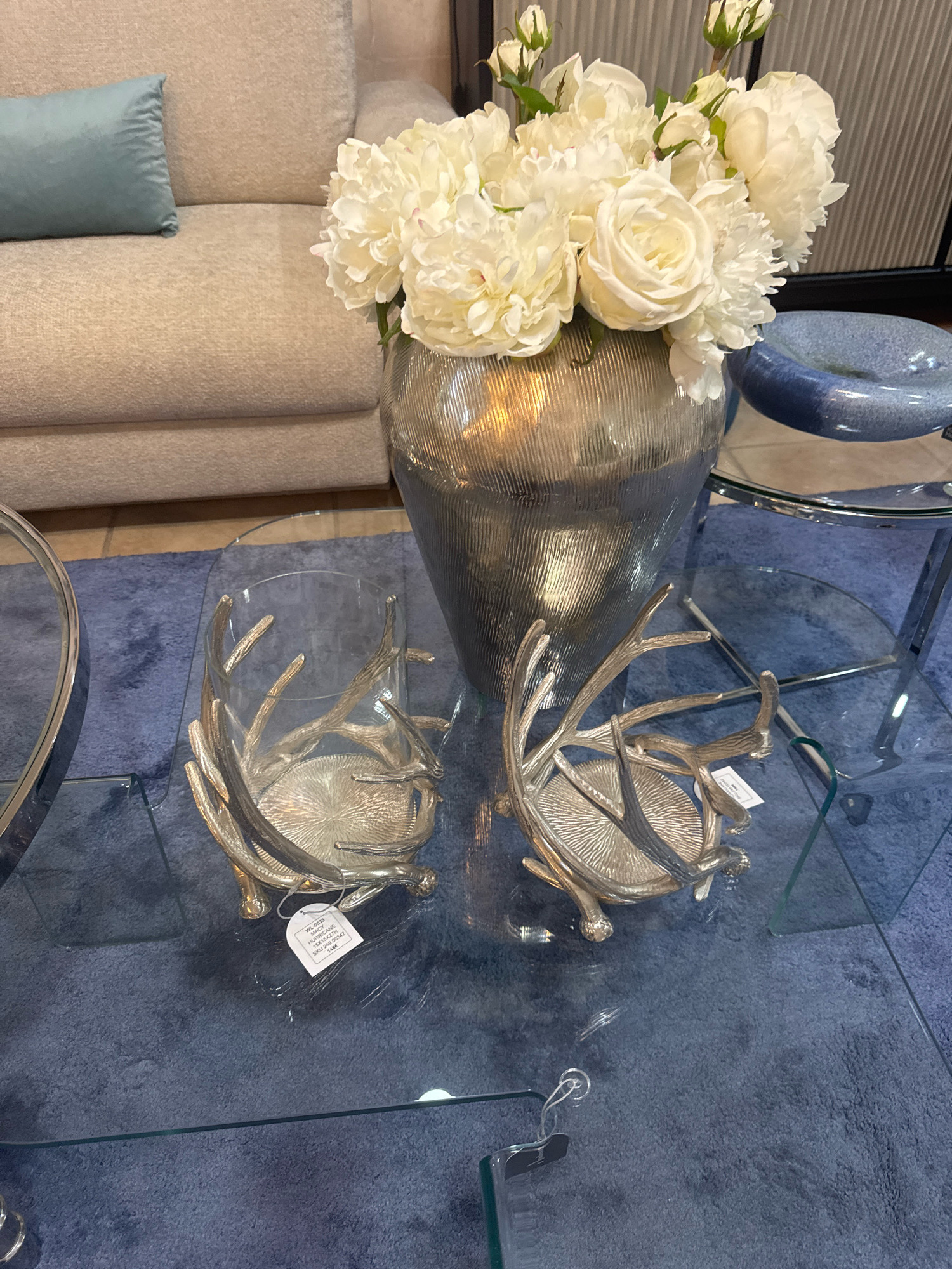 In a domestic interior setting, possibly a living room, a close-up view captures a glass-topped coffee table adorned with decorative items. The table rests on a textured blue rug. Prominently featured are two ornate, silver candle holders with branching, antler-like designs. To the left, a partially visible chrome-rimmed circular object, possibly another table or a tray, is present. Behind the candle holders, a tall, ribbed silver vase holds a generous bouquet of white peonies and roses. A light blue throw pillow rests on the arm of a beige upholstered sofa in the background. The scene suggests a staged display or an organized living space, with the focus on decorative accents. The setting is likely Agios Athanasios Municipality, Cyprus, based on the provided location context.