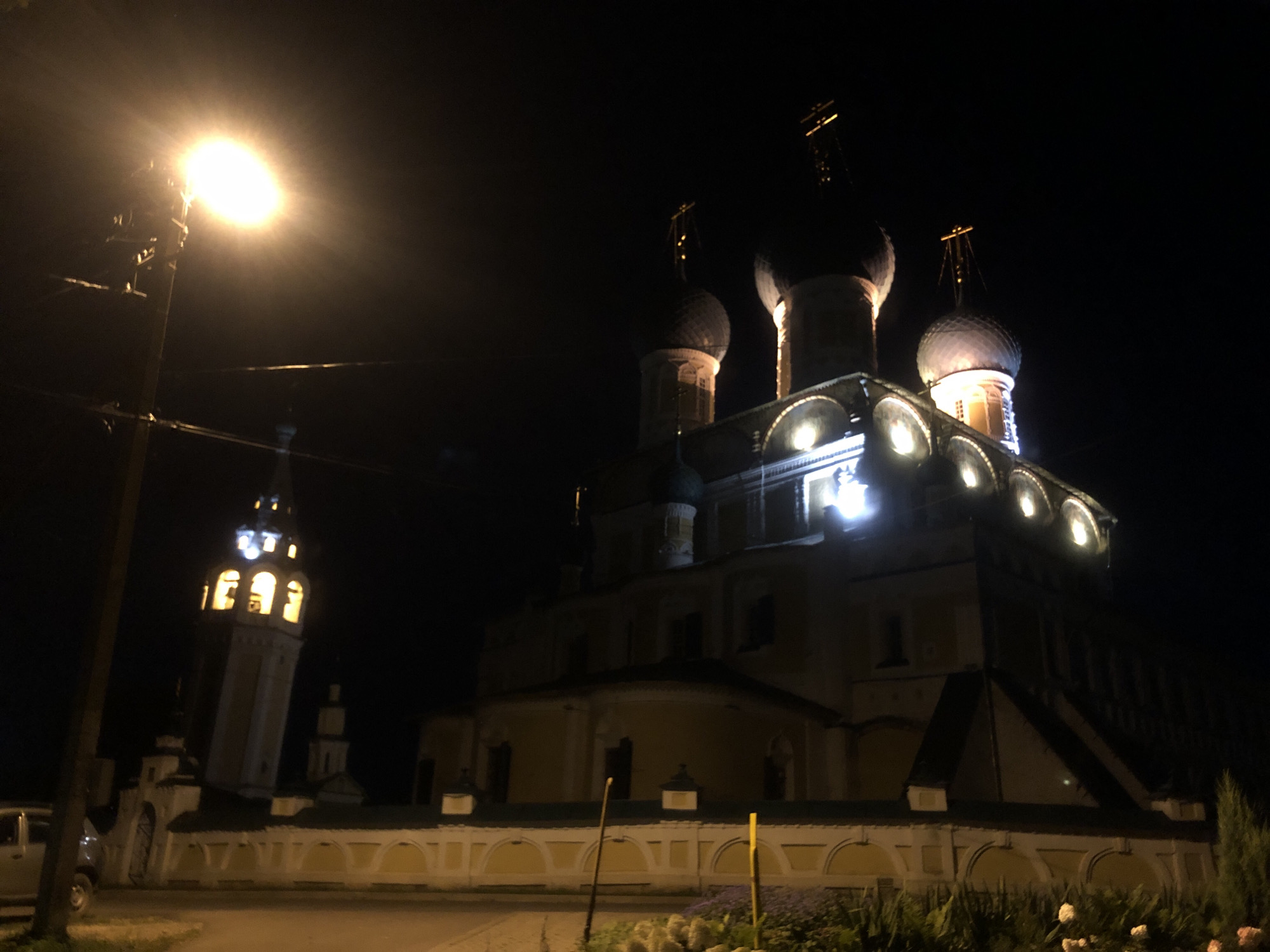 A low-angle, nighttime photograph captures the illuminated ensemble of the Resurrection Cathedral and bell tower in Tutaev, Russia, as seen from across a paved area. The Resurrection Cathedral is the dominant subject, with its multi-domed architecture and a towering bell tower to its left. Both structures are aglow with artificial lights, highlighting their ornate details against the dark sky. A street lamp on the left side of the frame emits a bright, warm light, casting a glow on a wooden utility pole and power lines. In the foreground, a white wall with arched alcoves stretches horizontally across the image, demarcating the boundary of the cathedral grounds. A dark-colored vehicle is partially visible on the left edge of the frame. The background is a deep black, characteristic of a night scene, with no visible stars or moon. The overall impression is one of serene, architectural grandeur bathed in artificial light.