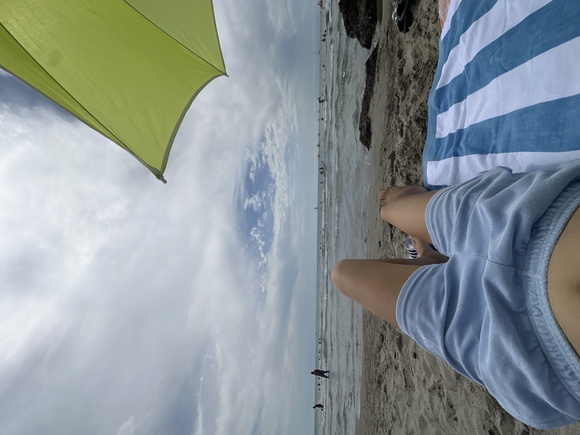 The image shows a first-person perspective of a person lying on a beach. The person is wearing light blue shorts and is lying on a blue and white striped towel. Their legs and feet are visible in the foreground. Behind them, the ocean stretches out to the horizon with several boats and people visible in the water. The sky is overcast with scattered clouds. A bright green umbrella is partially visible in the upper right corner of the frame. The scene appears to be a casual beach setting on a cloudy day.