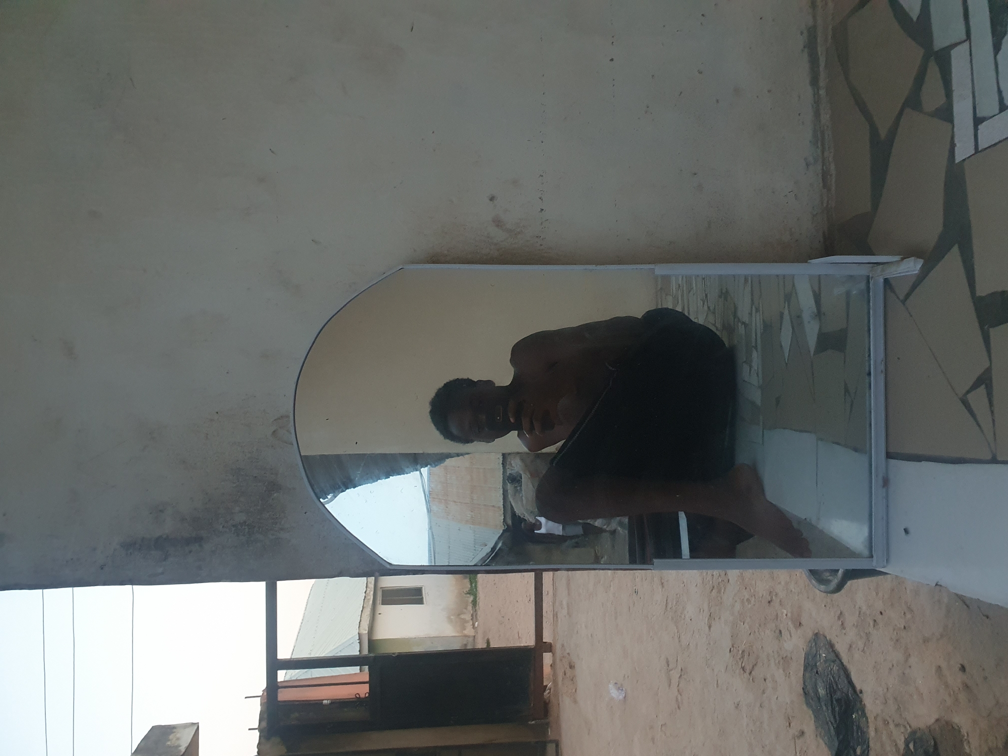 An outdoor, slightly rotated image captures a young man reflected in a mirror, presumably taking a selfie or being photographed. The man is shirtless, wearing dark bottoms, and is in a crouching or squatting position, looking towards the camera with a neutral expression.
The mirror, which has a simple frame, is positioned vertically against a wall. Its surface reflects not only the man but also an outdoor scene behind him, revealing residential buildings with light-colored walls and possibly metal roofs, suggesting a semi-urban or rural environment. A faint reflection of another person in a white sleeveless top is visible further back within the mirror's view.
Outside the mirror's frame, the immediate surroundings include a plain, somewhat weathered light-colored wall to the left (top of the image as presented) and a wall adorned with irregular, light-toned mosaic-like tiles to the right (bottom of the image). The ground in the foreground is visible as bare, sandy, and dusty earth with some debris. Beyond the mirror, in the background, more buildings and parts of a metal railing or structure are visible under a bright sky. The scene is brightly lit, indicating daytime with clear weather. There is no visible text in the image. The overall setting is consistent with an outdoor area in Lokoja, Nigeria.