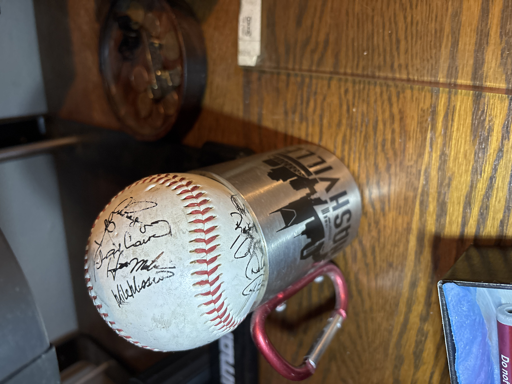 This is a medium shot, eye-level, indoor photograph of a baseball resting on a metallic mug. The baseball is white with red stitching and is covered in black ink signatures. The mug is silver and has a silhouette of a city skyline with the word "NASHVILLE" printed below it. A red carabiner is attached to the mug's handle. The objects are placed on a wooden surface, possibly a table. In the background, to the right of the mug, is a circular dish filled with coins. To the left of the baseball and mug, a portion of a black object with white lettering is visible. The overall lighting suggests an indoor environment, possibly a room with ambient light. There is no indication of the specific location within Round Rock, Texas.