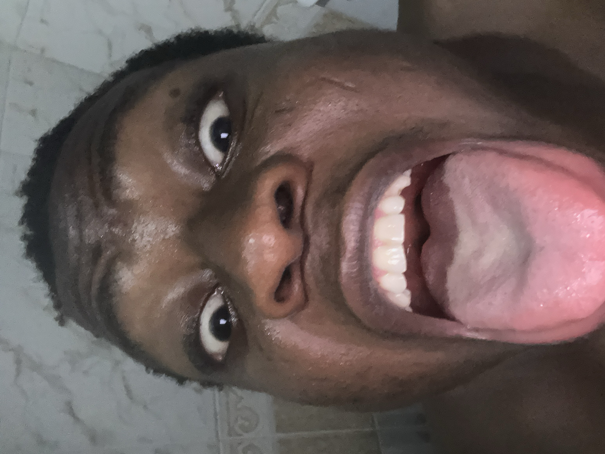 A close-up, slightly tilted perspective of a dark-skinned person's face with their mouth wide open and tongue protruding. The person has short, dark hair, and their eyes are wide open. Their teeth are visible inside their mouth. The background appears to be a tiled wall, possibly in a bathroom. The location is Unknown, Unknown.