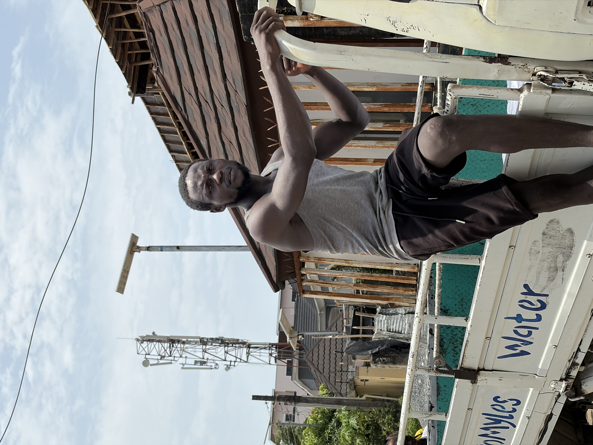 A man in a grey tank top and dark shorts is positioned on the side of a white truck with "Myles Water" written on it. He is gripping a white bar, appearing to climb onto or off the vehicle. The background includes buildings with corrugated metal roofing and a communication tower against a partly cloudy sky. The scene is located in Igboefon, Nigeria, suggested by the truck's branding "Myles Water."