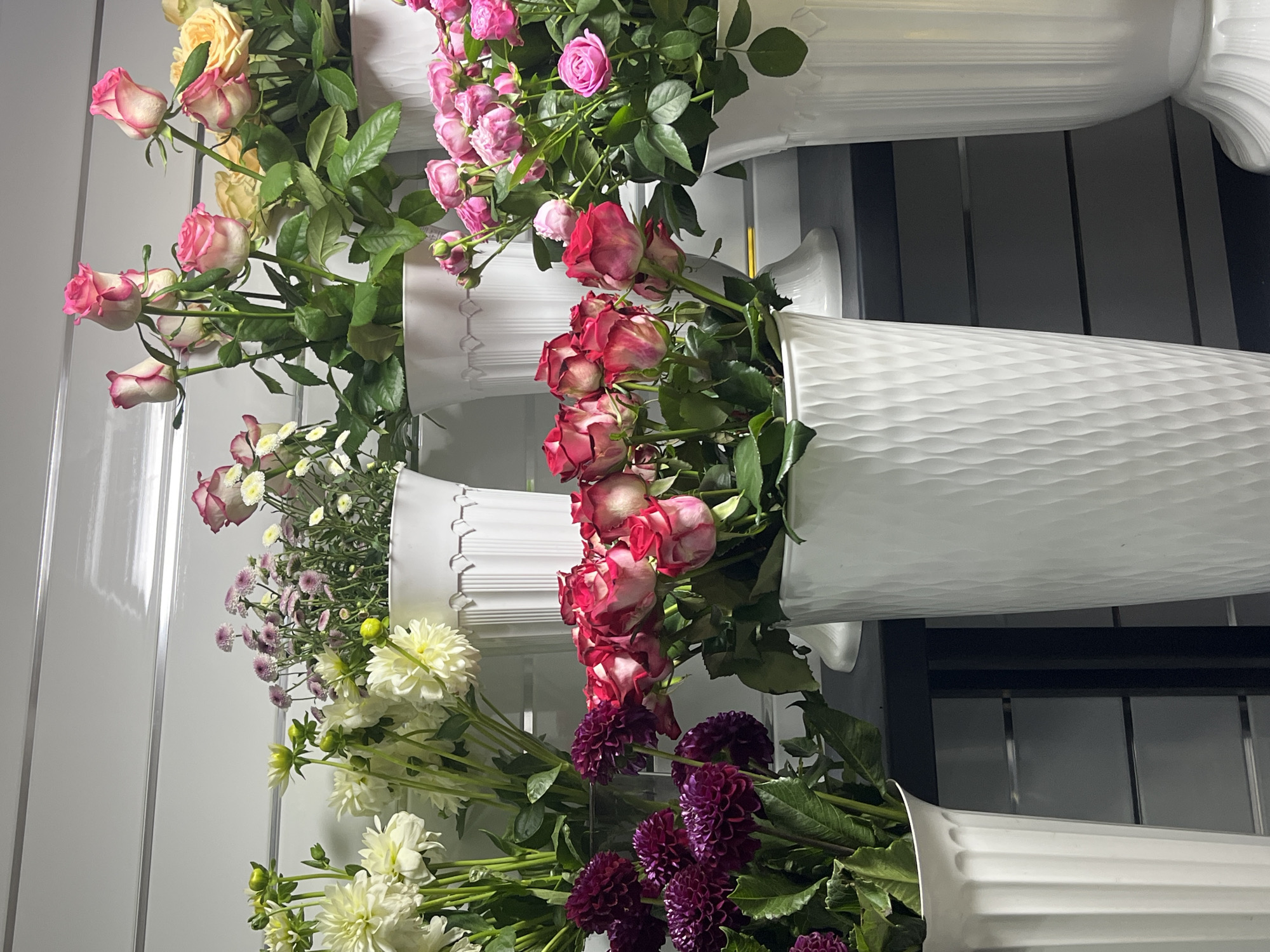 The image shows a close-up of a flower display in what appears to be a florist shop. There are several vases filled with various types of flowers. In the foreground, to the left, is a tall white vase containing a bouquet of deep purple dahlia-like flowers with dark green leaves. To the right of this vase is a larger, textured white vase filled with red and white roses. Further back, a large white vase holds a bouquet of pink and white roses, and behind that, another white vase contains yellow roses and small pink and white flowers. The background is a light-colored wall with horizontal grooves. The lighting suggests an indoor setting. There are no people visible in the image.