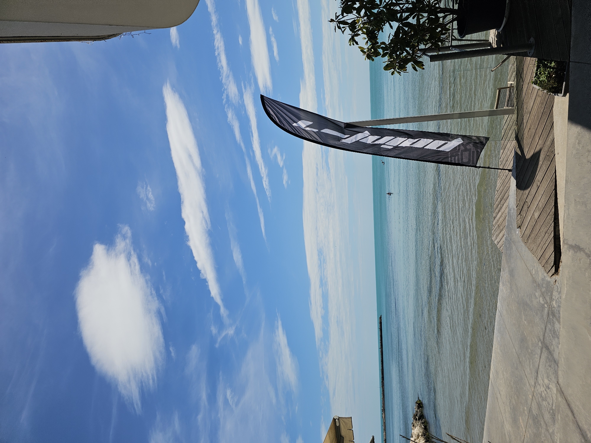 The image shows a coastal scene with a blue sky populated by scattered white clouds. An elongated black flag with white lettering is positioned near the foreground, next to a wooden deck. The ocean stretches toward the horizon with a light turquoise hue. Vegetation is visible near the top right of the image. The location is the city of Unknown, Unknown.