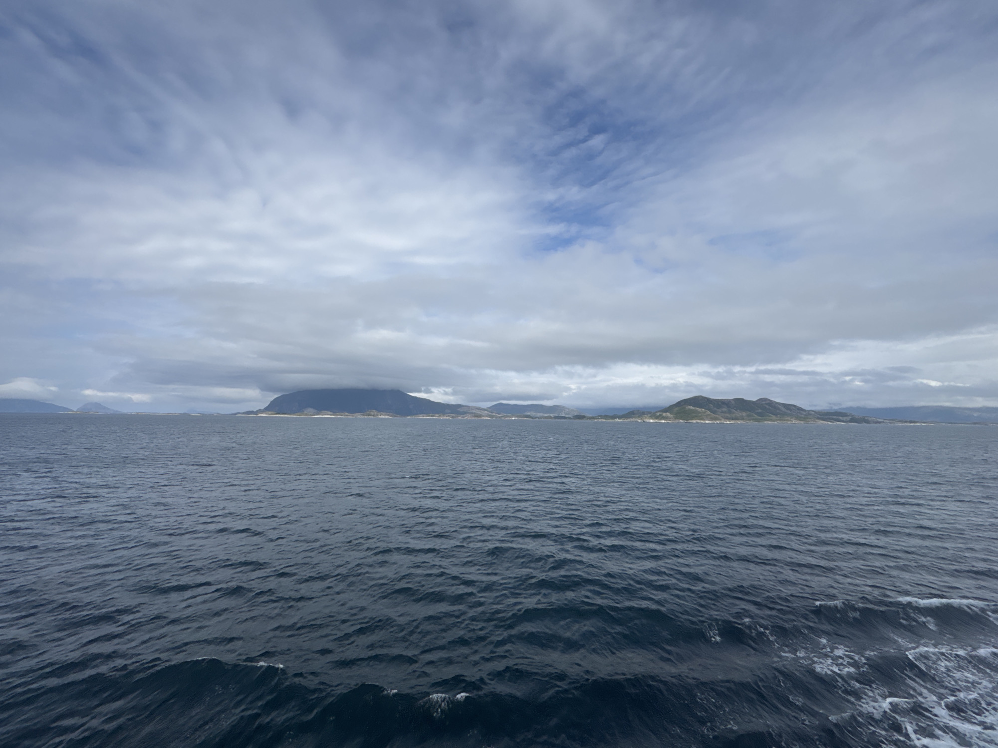 A wide shot captures a body of water with islands on the horizon under a cloudy sky. The water is dark blue with small waves creating white foam and crests in the foreground. Distant islands are visible, with some appearing mountainous and others lower and covered in vegetation. The sky is filled with a mixture of light and dark clouds, with patches of blue sky showing through. The scene appears to be a coastal landscape in Onøya, Norway. No people or specific objects are identifiable in the image.
