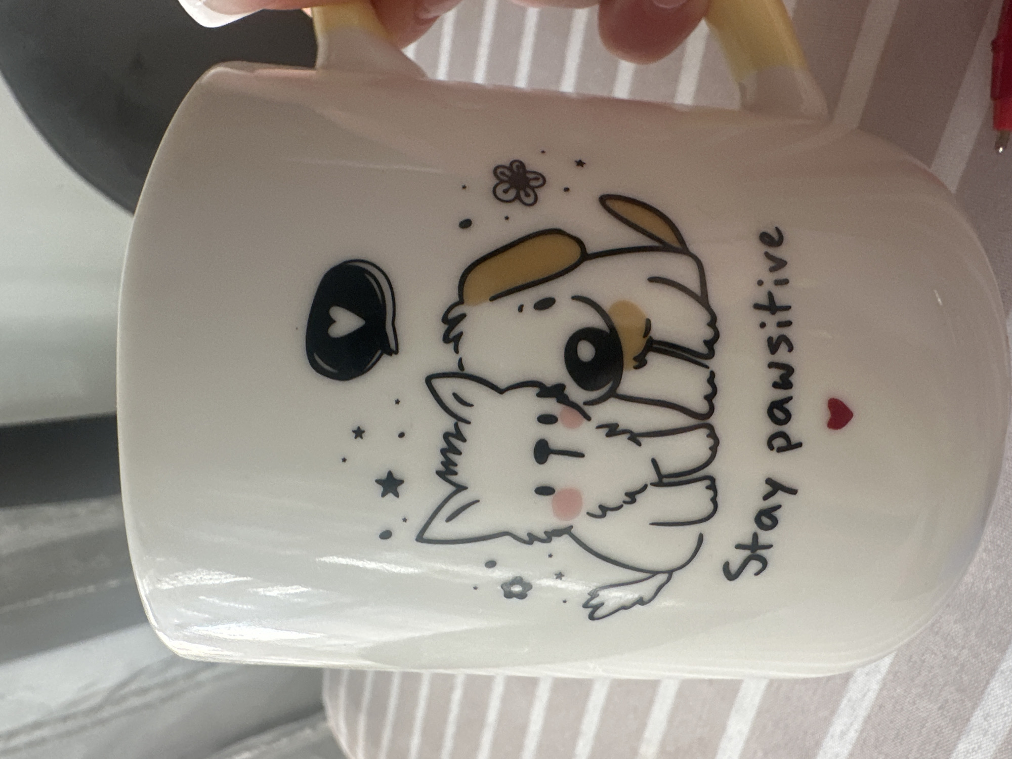 The image shows a white ceramic mug with a cartoon illustration of two puppies cuddling. One puppy has tan ears and a black dot for an eye. The other puppy is white with pink cheeks. A black speech bubble with a white heart is to the left of the puppies. The words "Stay pawsitive" are printed in black lettering on the right side of the mug with a small red heart serving as the dot over the "i" in "pawsitive." The mug has a yellow handle. The background appears to be a striped fabric surface. There is no indication of the location being in Sofia, Bulgaria.