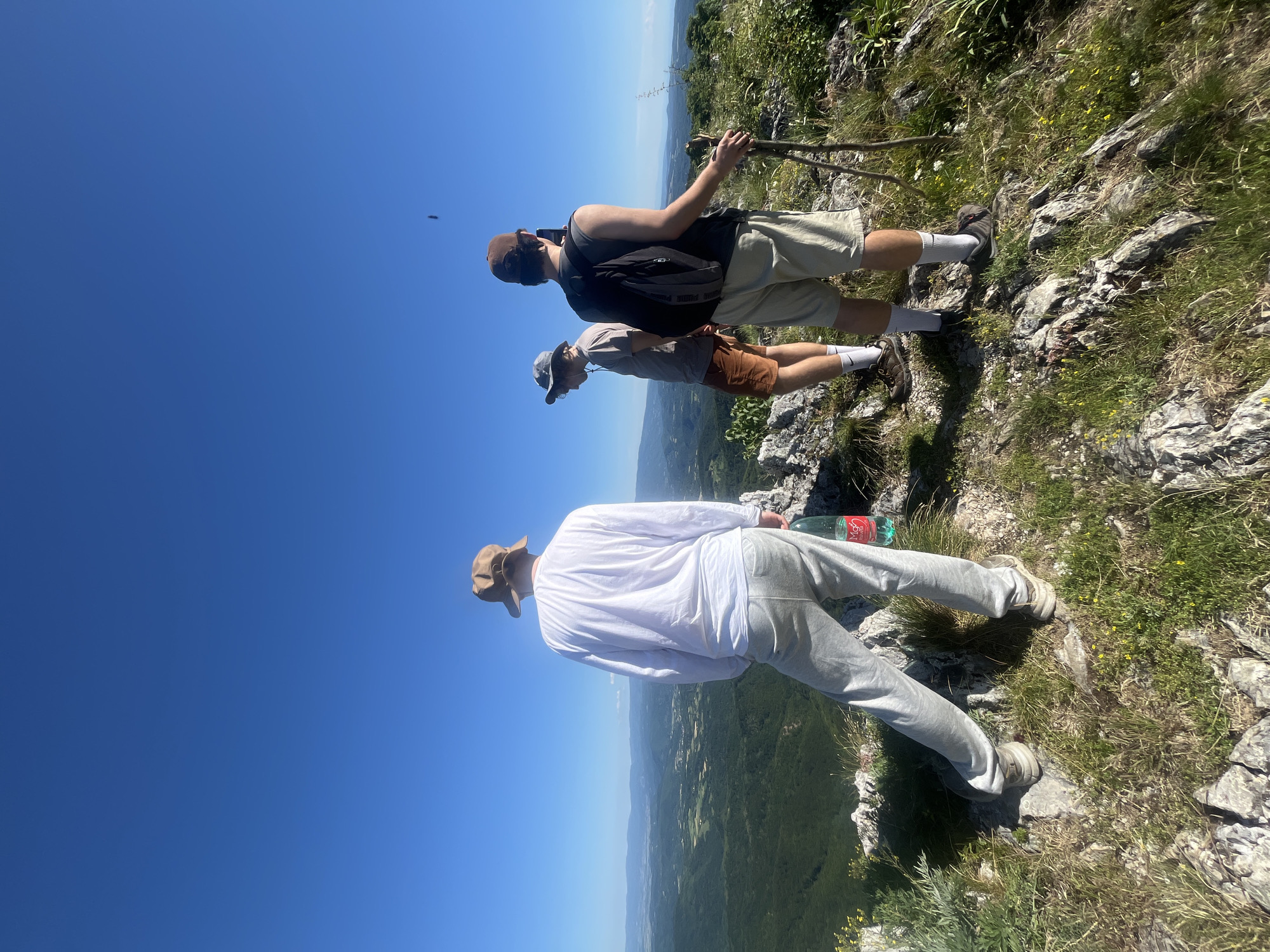 Three individuals are standing on a rocky, grassy precipice overlooking a valley with rolling, forested hills under a clear blue sky. The person on the left, viewed from the back, wears a tan bucket hat, a long-sleeved white shirt, and grey sweatpants. A green plastic bottle of water is held in their right hand. In the middle, another individual, facing left and slightly away from the camera, wears a grey bucket hat, a grey t-shirt, and orange shorts. The person on the right, facing away from the camera and holding two wooden hiking poles, wears a brown baseball cap, a dark tank top over a grey t-shirt, and light beige shorts. They are holding a mobile phone in their right hand, appearing to take a photo or video. The environment is natural and mountainous, with sparse vegetation on the rocky terrain. The scene suggests a hiking or outdoor exploration activity.