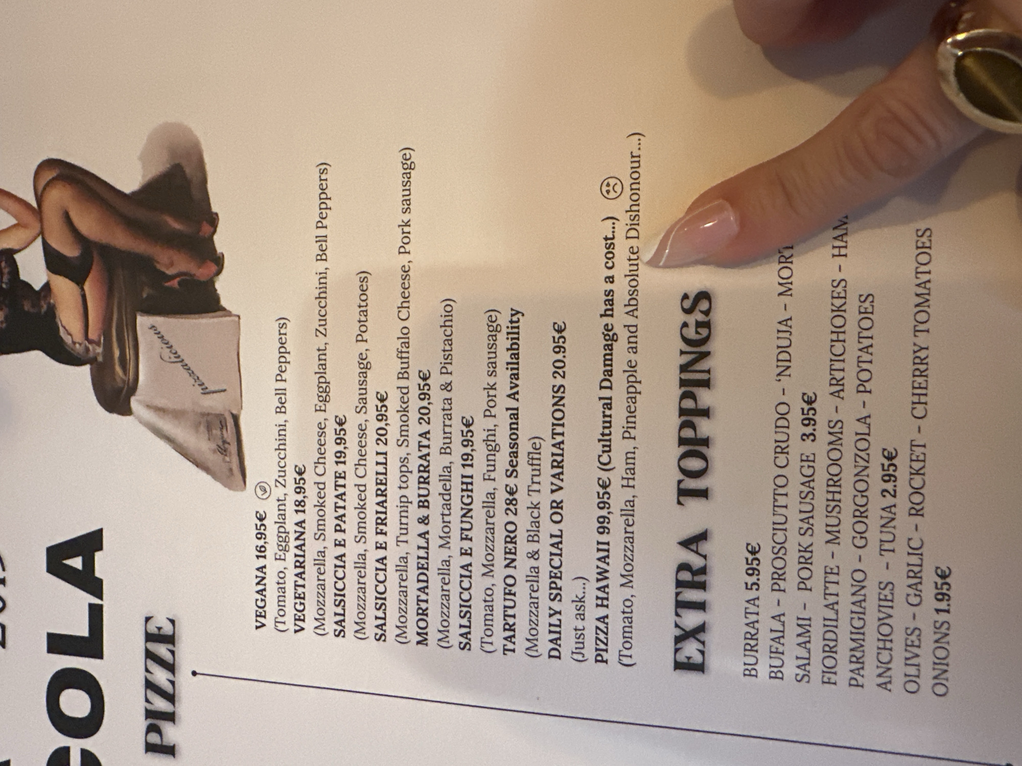 This is a close-up, high-angle shot of a menu with text and a hand pointing to it. The menu appears to be for pizza and features a variety of topping options and prices. The text is in Italian and English. To the left of the menu, a portion of an image is visible, depicting a stylized drawing of a person in a provocative pose. The background is a light color, possibly a wall or table. The overall impression is a casual dining setting. The image has a warm, indoor lighting. There is no indication of weather or time of day. The hand pointing at the menu has manicured nails, suggesting a person is currently interacting with the menu.