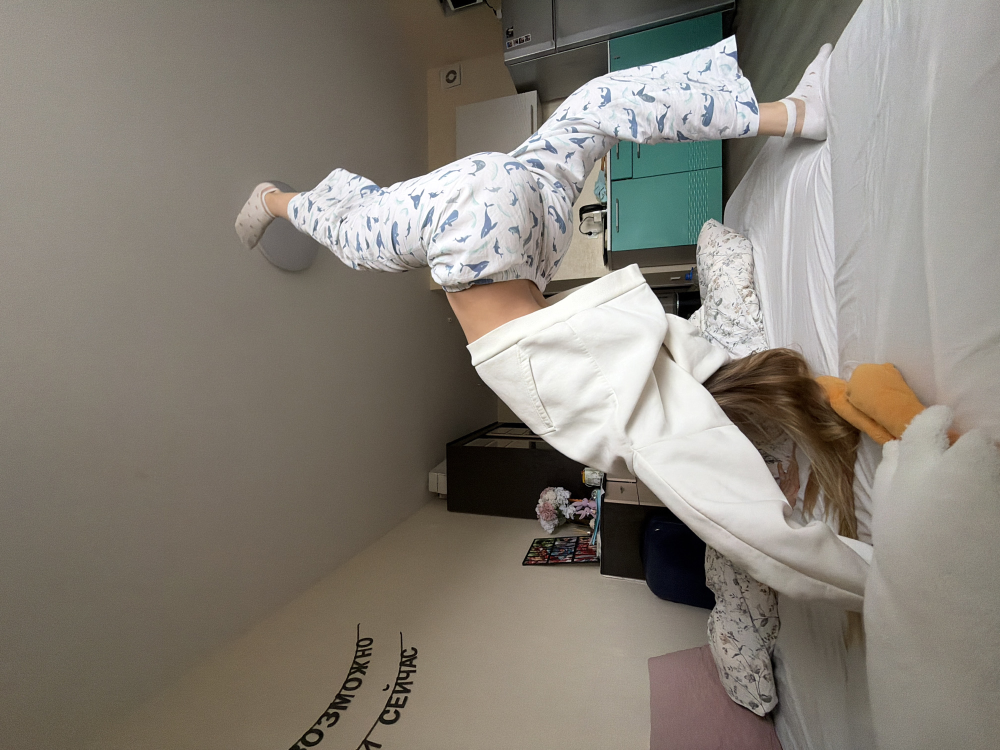 A young woman in a white hoodie and whale-print pajamas is upside down, balancing with her feet against a wall and the edge of a bed. The location is a bedroom in Izhevsk, Russia, based on the Cyrillic text on the wall. The room includes a bed with white sheets, various cushions, bedside furniture, and a rug with flowers.
