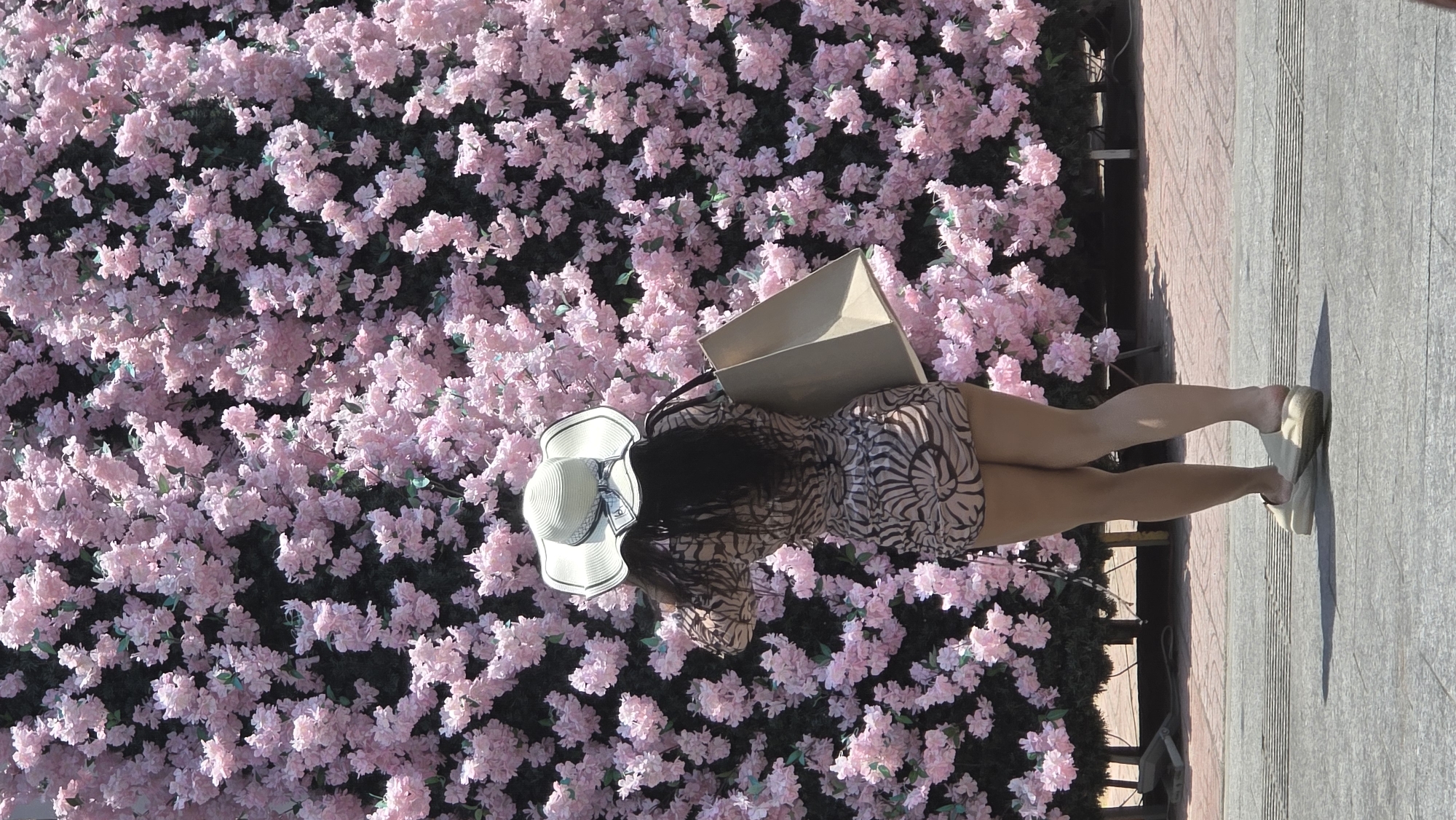 A young woman is standing next to a tree or wall covered in pink flowers. She is wearing a light-colored dress, a hat, and light-colored shoes. She is also holding a shopping bag. She is standing on a sidewalk with light-colored pavement. The location is potentially in Nha Trang, Vietnam, as specified in the prompt.