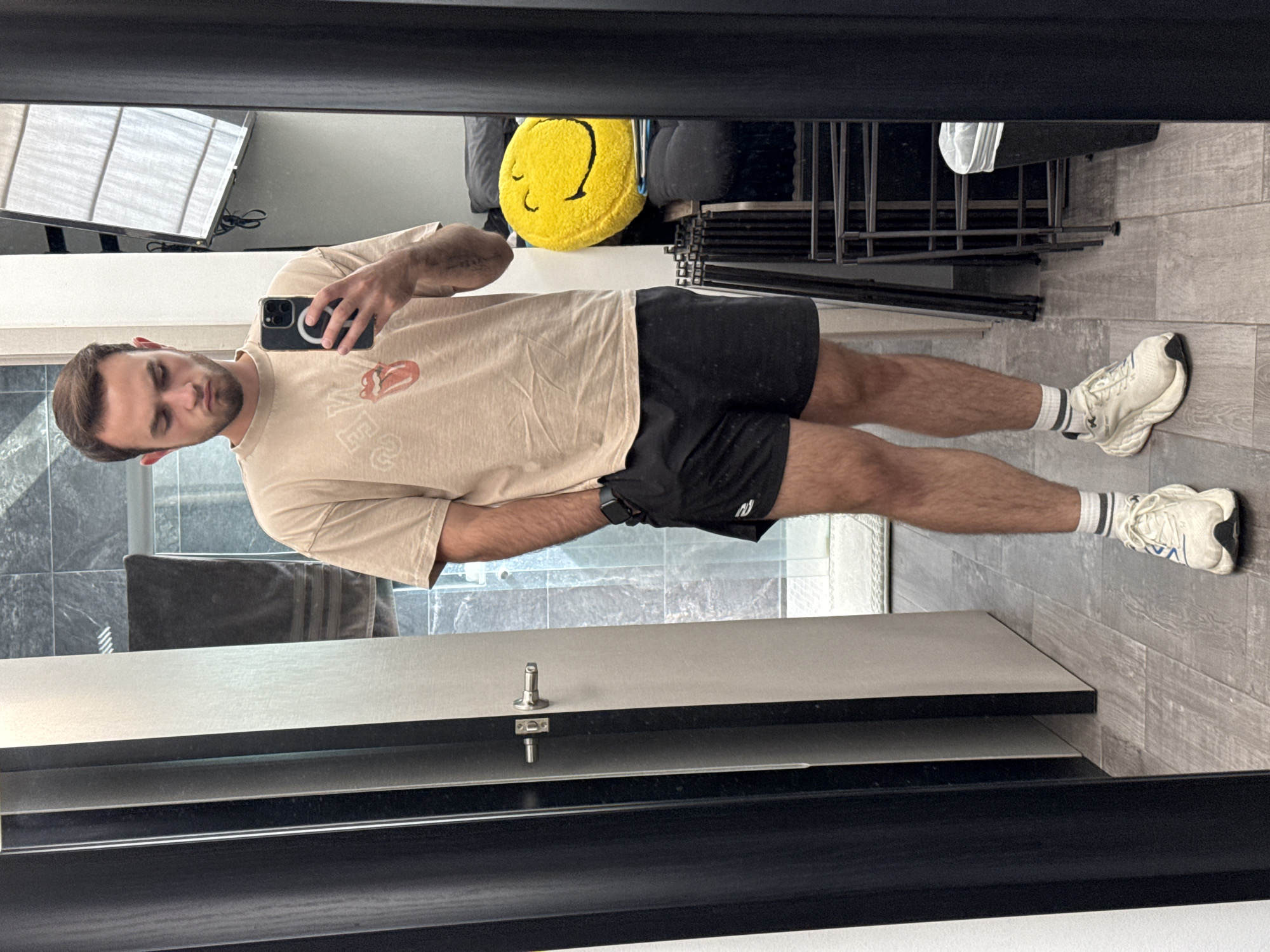 A man is taking a selfie in a bathroom mirror. He is wearing a beige t-shirt with a graphic, black athletic shorts, white socks with black stripes, and white sneakers. He is holding a black smartphone in his left hand. The bathroom has gray tile flooring and a vanity with a gray countertop and black cabinets. In the background, there is a window with blinds, a yellow smiley face pillow on top of stacked folding chairs, and a gray towel hanging near the mirror. The location in San Bernardino Tlaxcalancingo, Mexico is not discernible from the image.