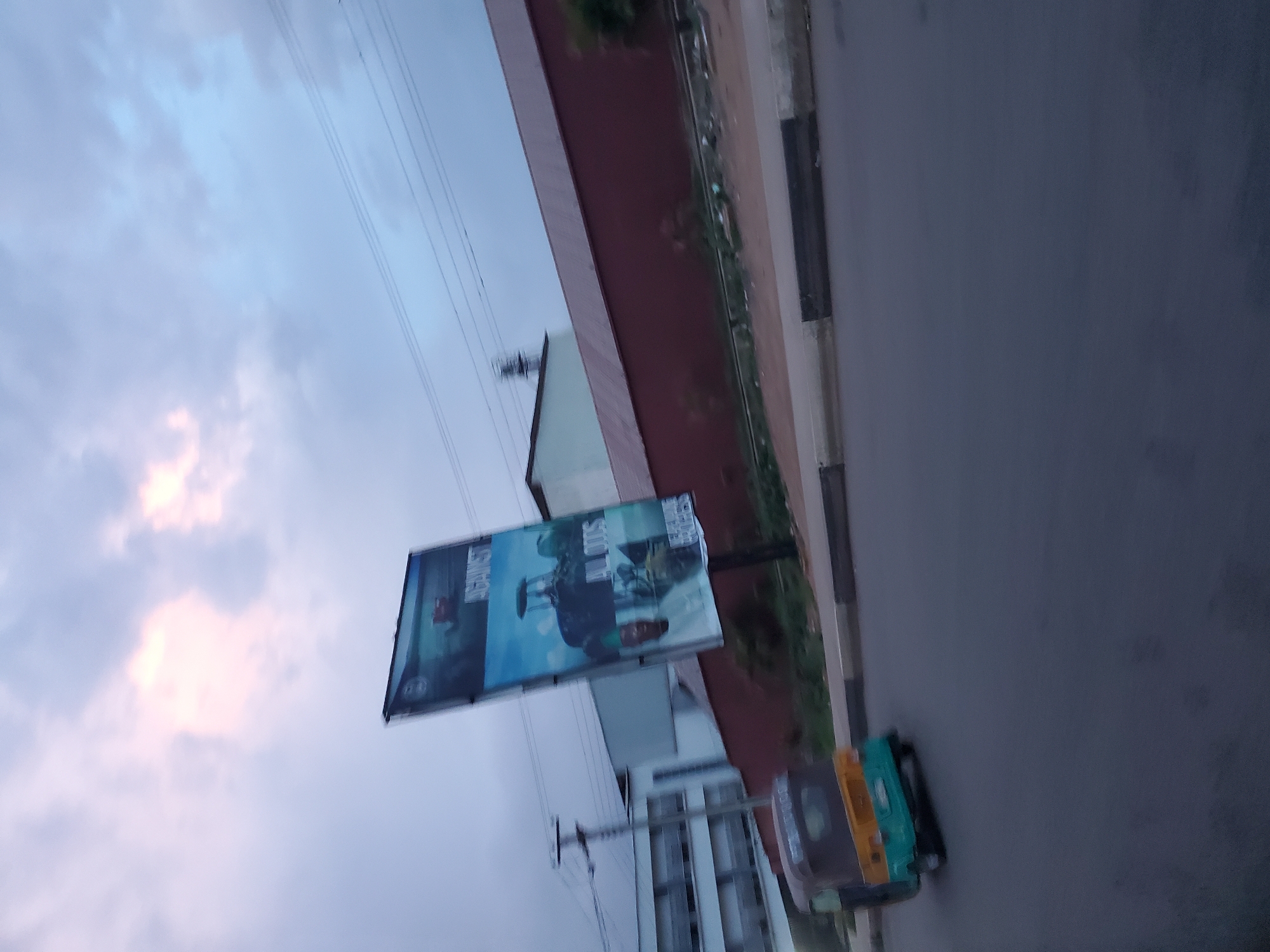 The image, taken in Jos, Nigeria, captures a street scene with a billboard in the background advertising agricultural machinery. A three-wheeled vehicle, commonly known as an "okada" or "keke," is prominently featured in the foreground, driving on an asphalt road. The billboard displays images of tractors and possibly other farming equipment, along with text that appears to promote agricultural solutions or products. The sky is overcast with soft, diffused light, suggesting either early morning or late afternoon. The surrounding environment includes buildings and a red-colored wall, typical of urban landscapes in Nigeria. The overall impression is that of a typical day in a Nigerian city, with elements of transportation and commerce visible.