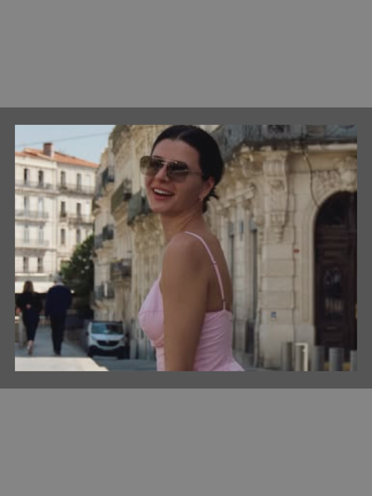 A woman is in the foreground, looking over her shoulder and smiling. She has dark hair and is wearing sunglasses and a pink spaghetti strap dress. In the background, two people are walking away from the camera on a street with buildings on either side. A white car is parked on the side of the street. The buildings appear to be in a European style with ornate details. The lighting suggests it is daytime.
