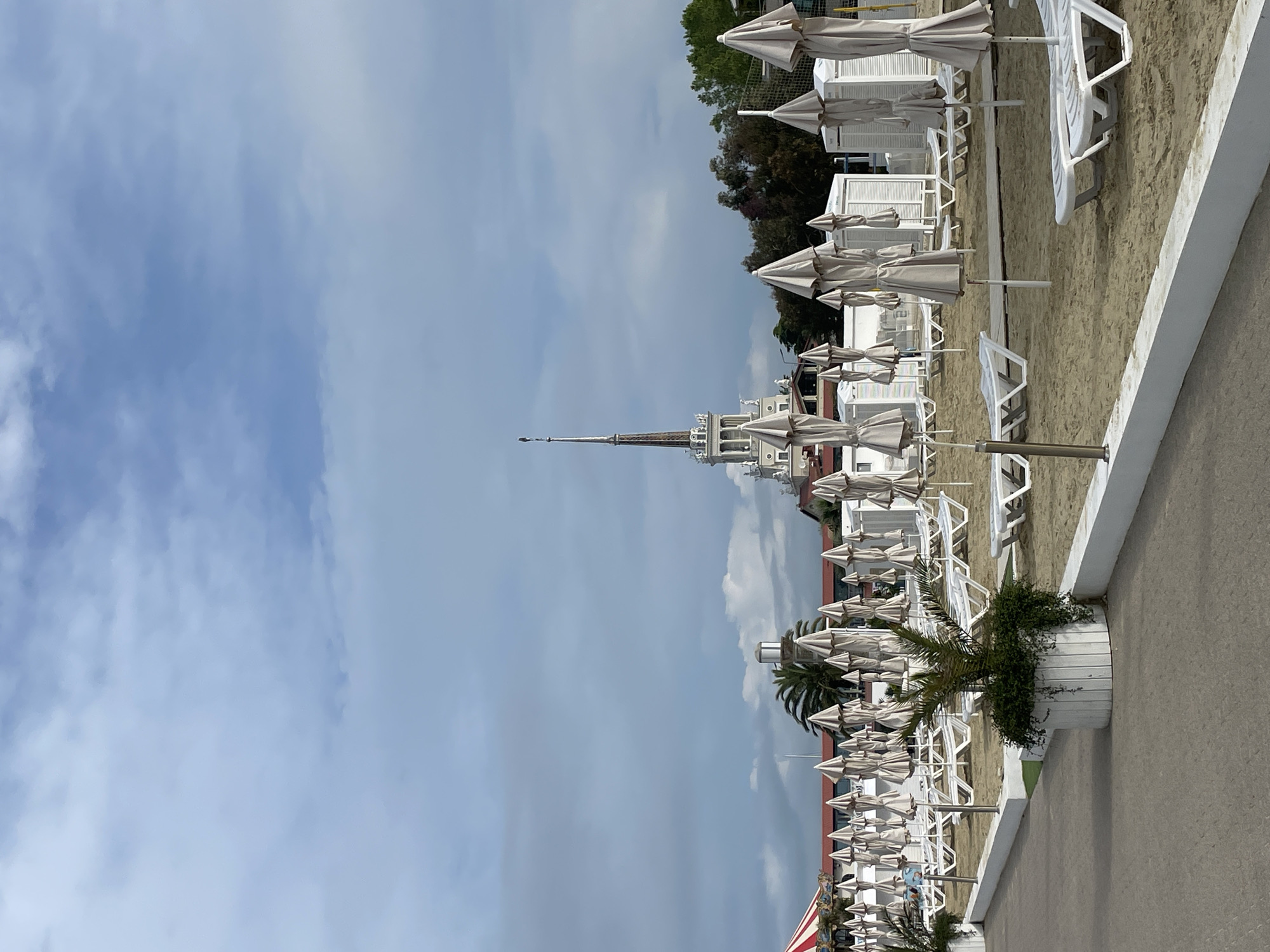 The image captures a beach scene with rows of closed umbrellas and white lounge chairs along the shore. In the background, there is a building with a prominent spire, which identifies the location as the beach in Sochi, Russia, near the Morskoi Vokzal (Sea Port). The sky is partially cloudy, and the image is oriented in a vertical format.