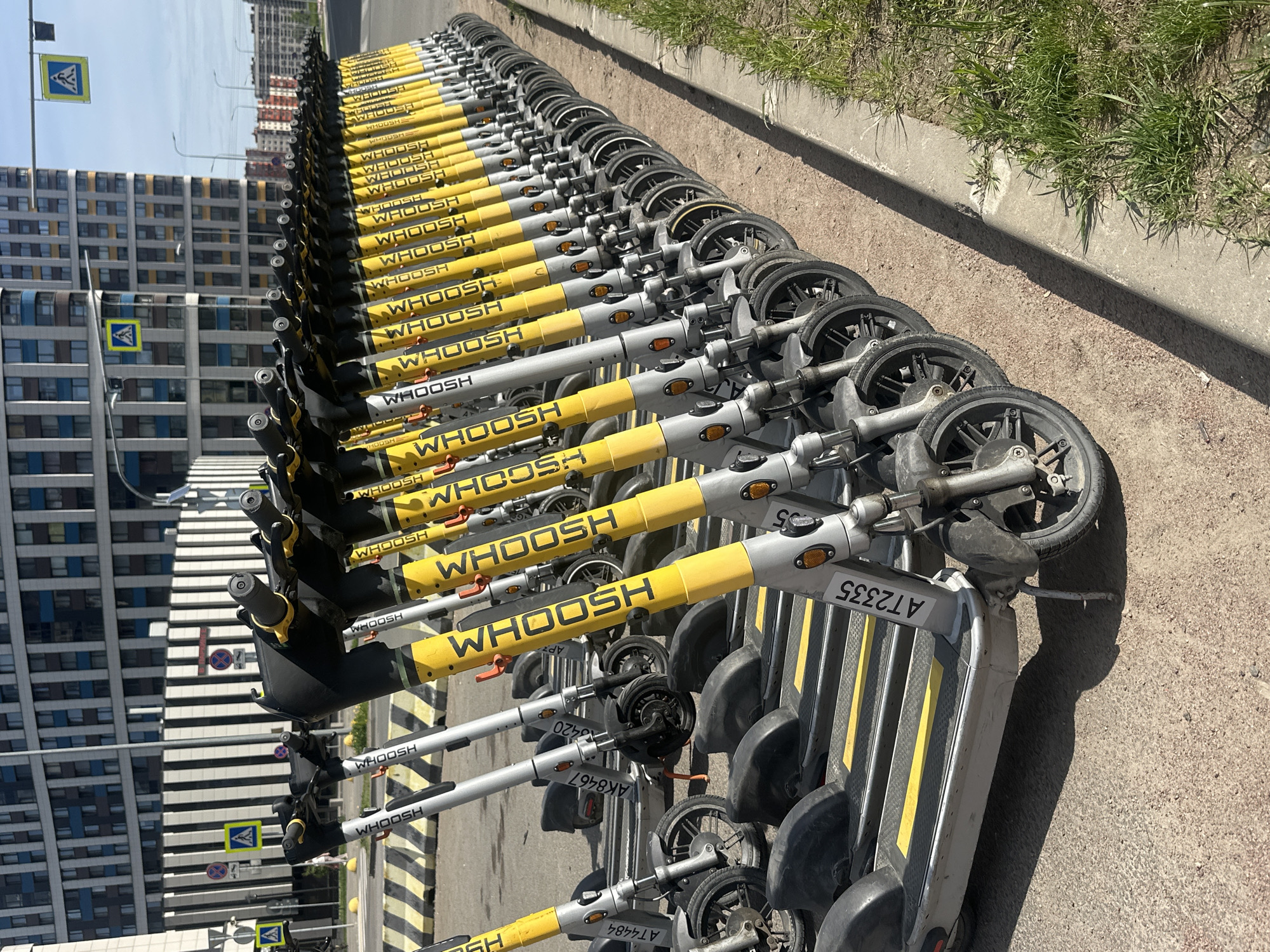 The image depicts a long row of electric scooters, likely belonging to a rental service, parked on a paved surface in Murino, Russia. The scooters are primarily gray with yellow accents and have the brand name "WHOOSH" prominently displayed. The scooters are lined up neatly, facing the same direction. A curb with grass is visible to the right. A multi-story building is on the left side of the image. A pedestrian crossing road sign is present. Scooter's registration plates are visible, with number AT2335 on the first scooter. The scene is well-lit.