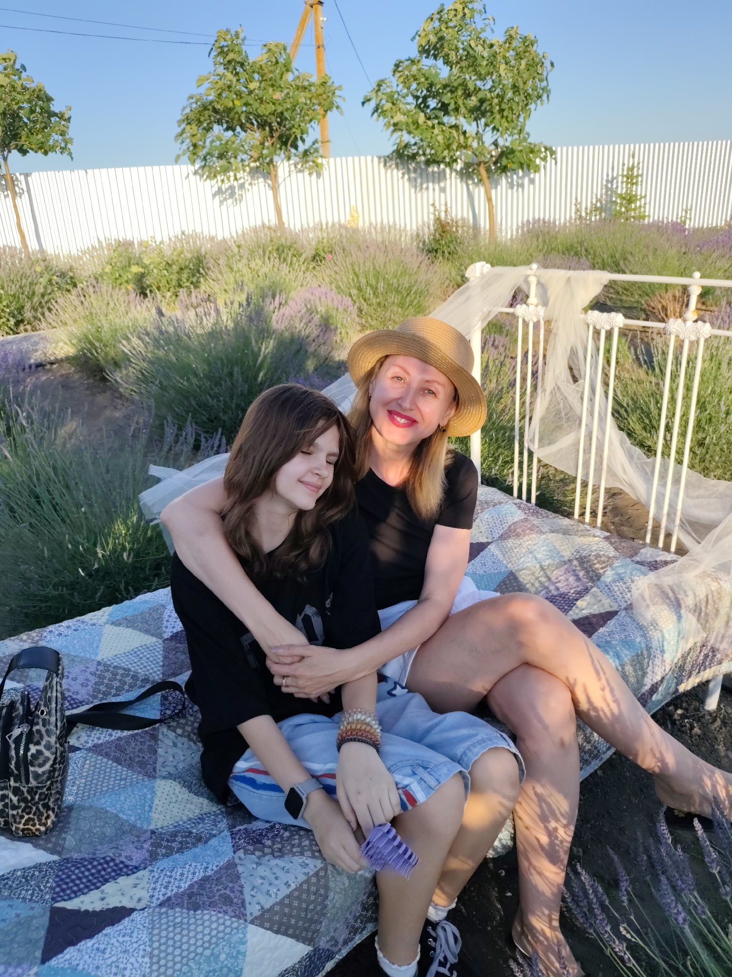 A woman and a teenage girl are sitting on a blanket spread on the ground, with a white metal bed frame behind them. The woman, wearing a straw hat, a black t-shirt, and white shorts, has her arm around the girl. The girl, with her eyes closed and a slight smile, is wearing a black t-shirt and blue denim shorts. They are in a field of lavender, with trees and a white fence in the background. The sun is setting, casting long shadows and creating a warm, golden light. The overall mood is one of affection and relaxation, possibly during a summer evening in Nechaiane, Ukraine. A leopard-print bag and bracelets on the girl's wrist are visible.