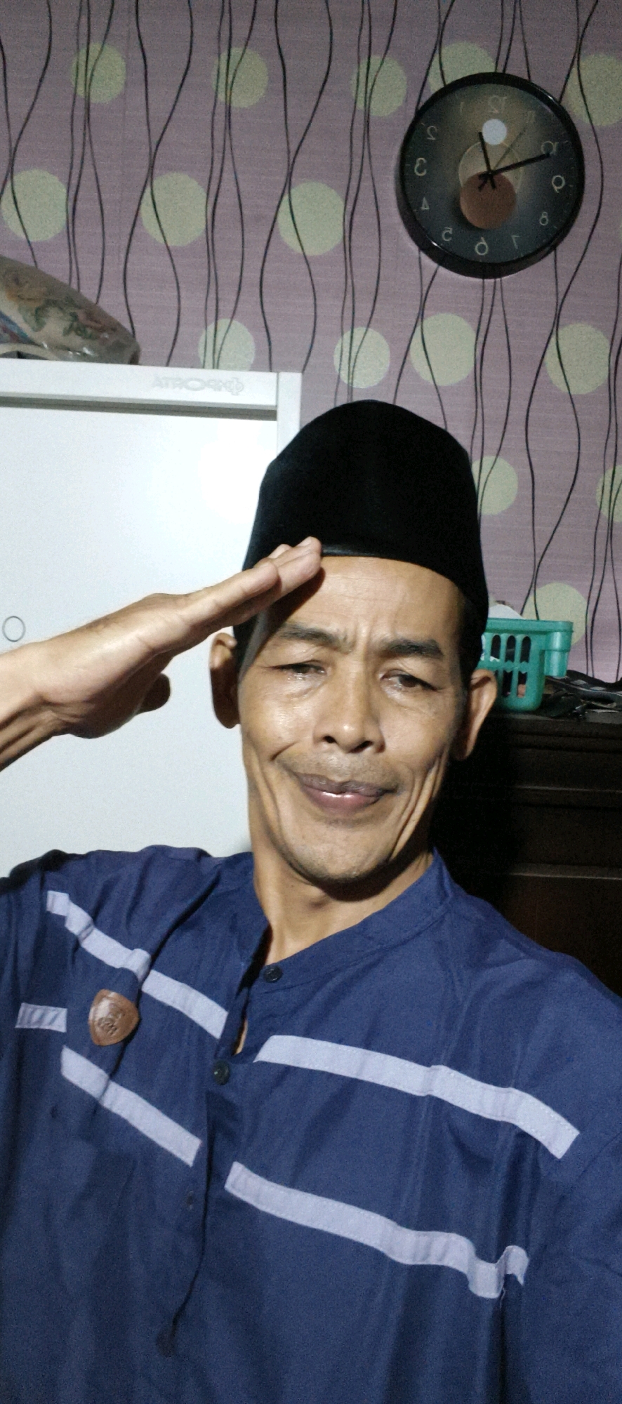 A man, identified as Indonesian, is captured in a medium close-up selfie. He is wearing a dark blue, collared shirt adorned with two light gray horizontal stripes across the chest. A black peci (cap) covers his head, and a brown emblem is affixed to the left side of his shirt. His right hand is raised in a saluting gesture, touching his forehead. The man's facial expression is a broad smile, with his mouth slightly open, revealing his teeth and tongue.
The background features a wall with purple wallpaper patterned with black wavy lines and pale yellow circles. To the upper right, a round black clock with a dark gray face and white numerals is mounted on the wall, showing the time as approximately 2:37. To the left of the clock, a white appliance, possibly a refrigerator, is partially visible. Further to the right, a dark wooden shelf holds a green plastic basket. The lighting appears to be artificial and possibly from an overhead source, casting shadows and highlighting the man's features. The scene is consistent with an indoor domestic setting in Muaro Jambi, Indonesia.